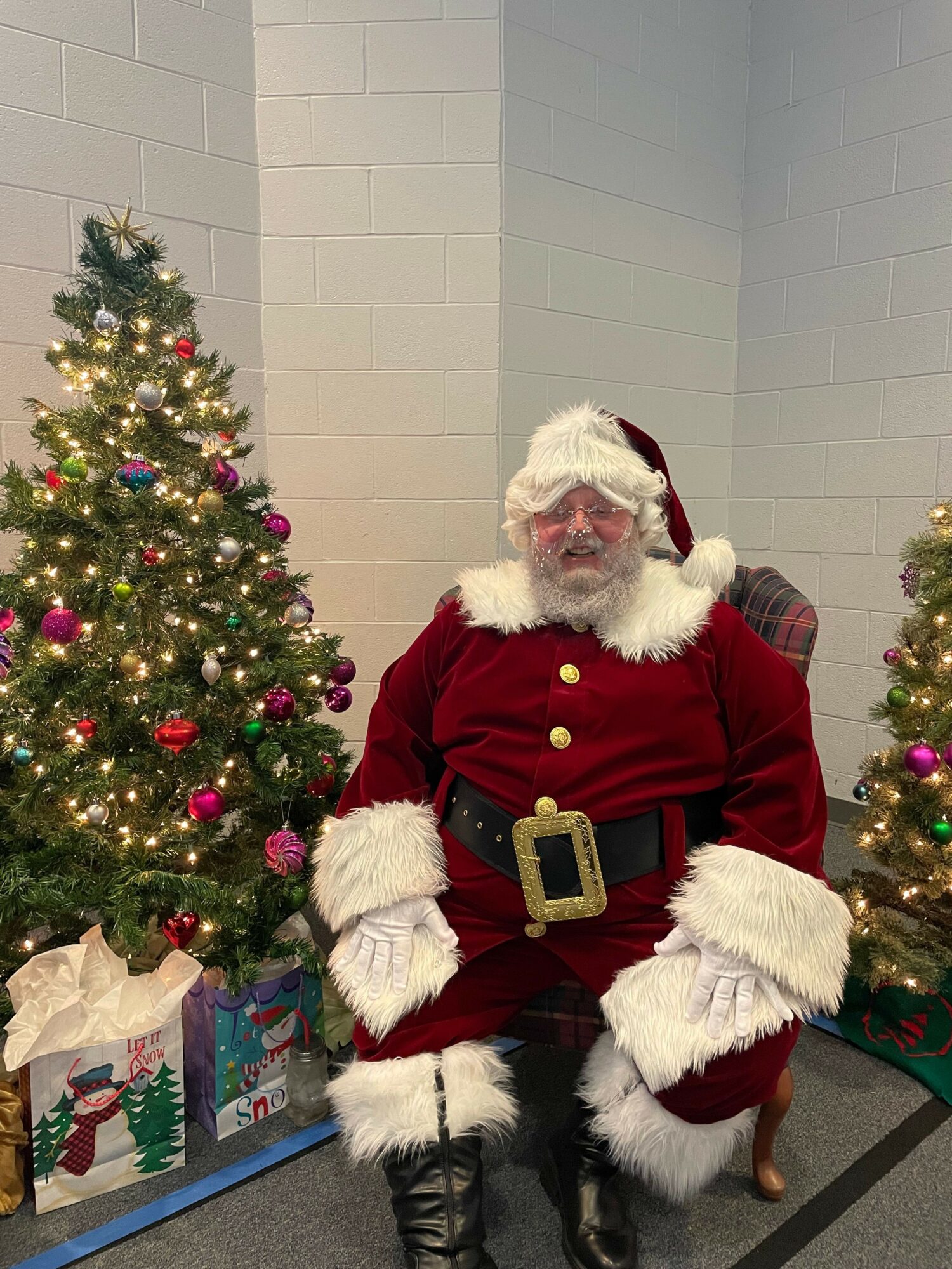 Person dressed as Santa Claus standing next to decorated Christmas tree with presents underneath, in a room with white walls.
