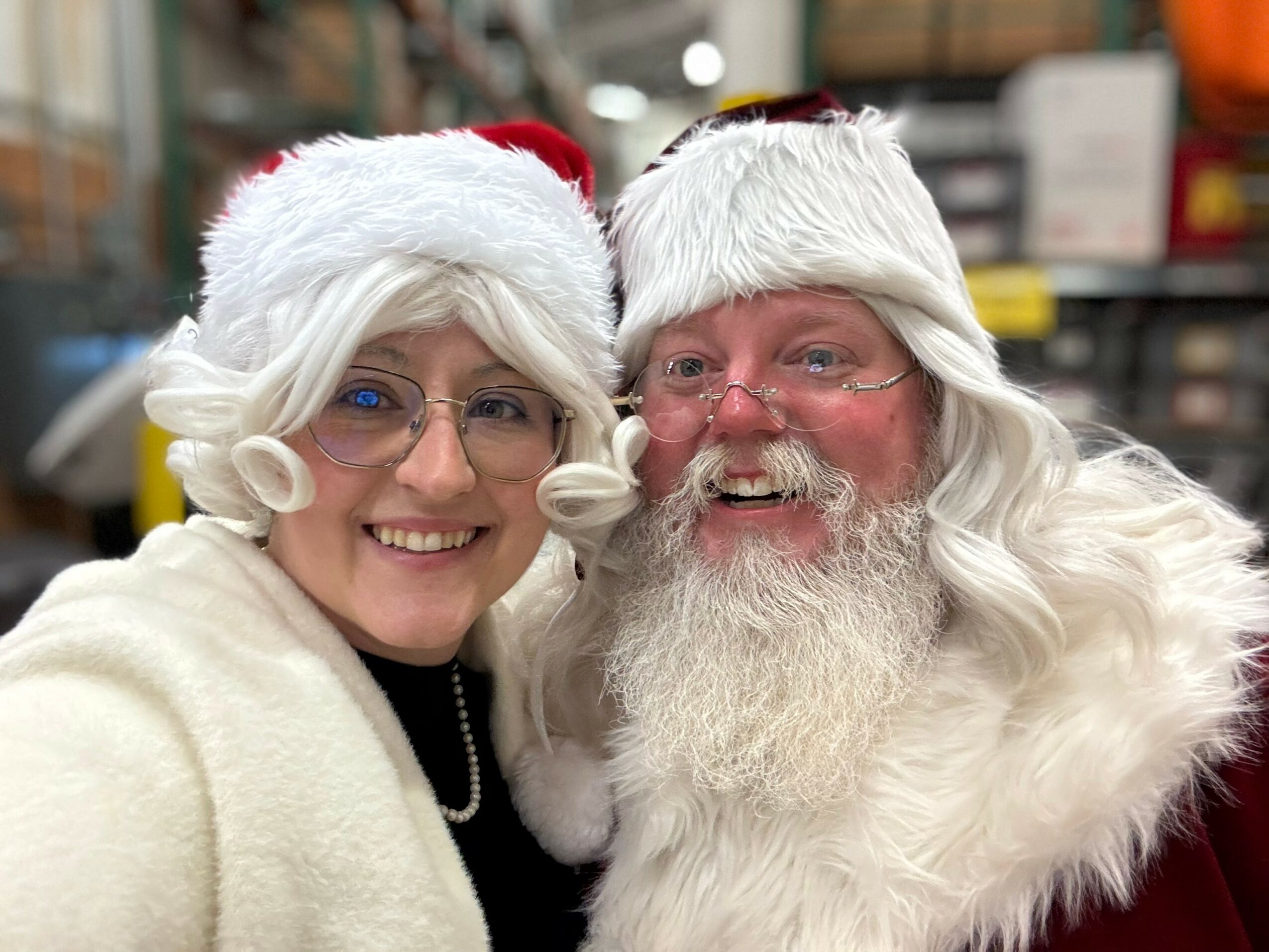 Two people dressed as Santa Claus and Mrs. Claus smiling, wearing red and white hats, in a festive setting.