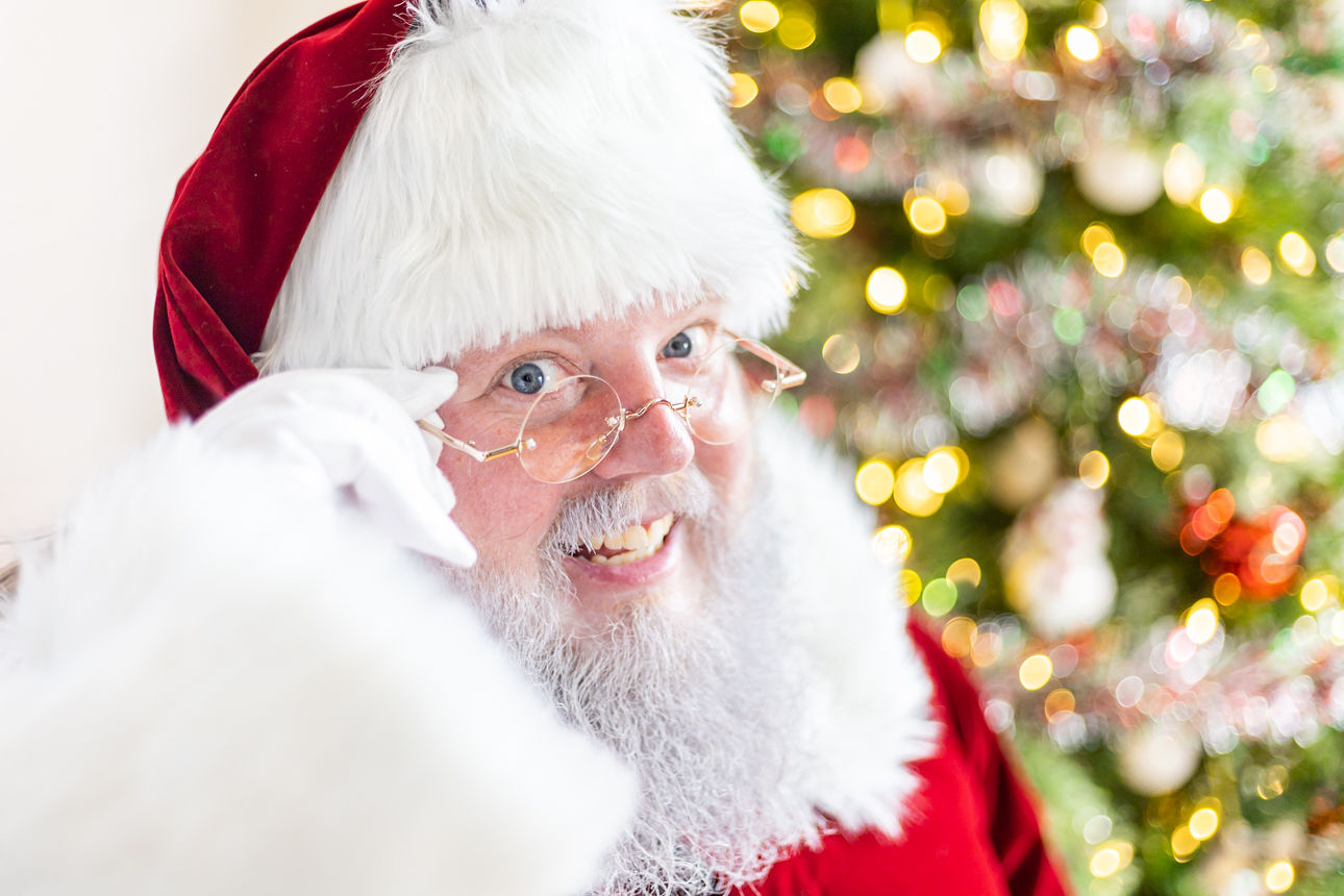 Santa Claus with glasses and a white beard, wearing a red hat, smiling near a decorated Christmas tree.