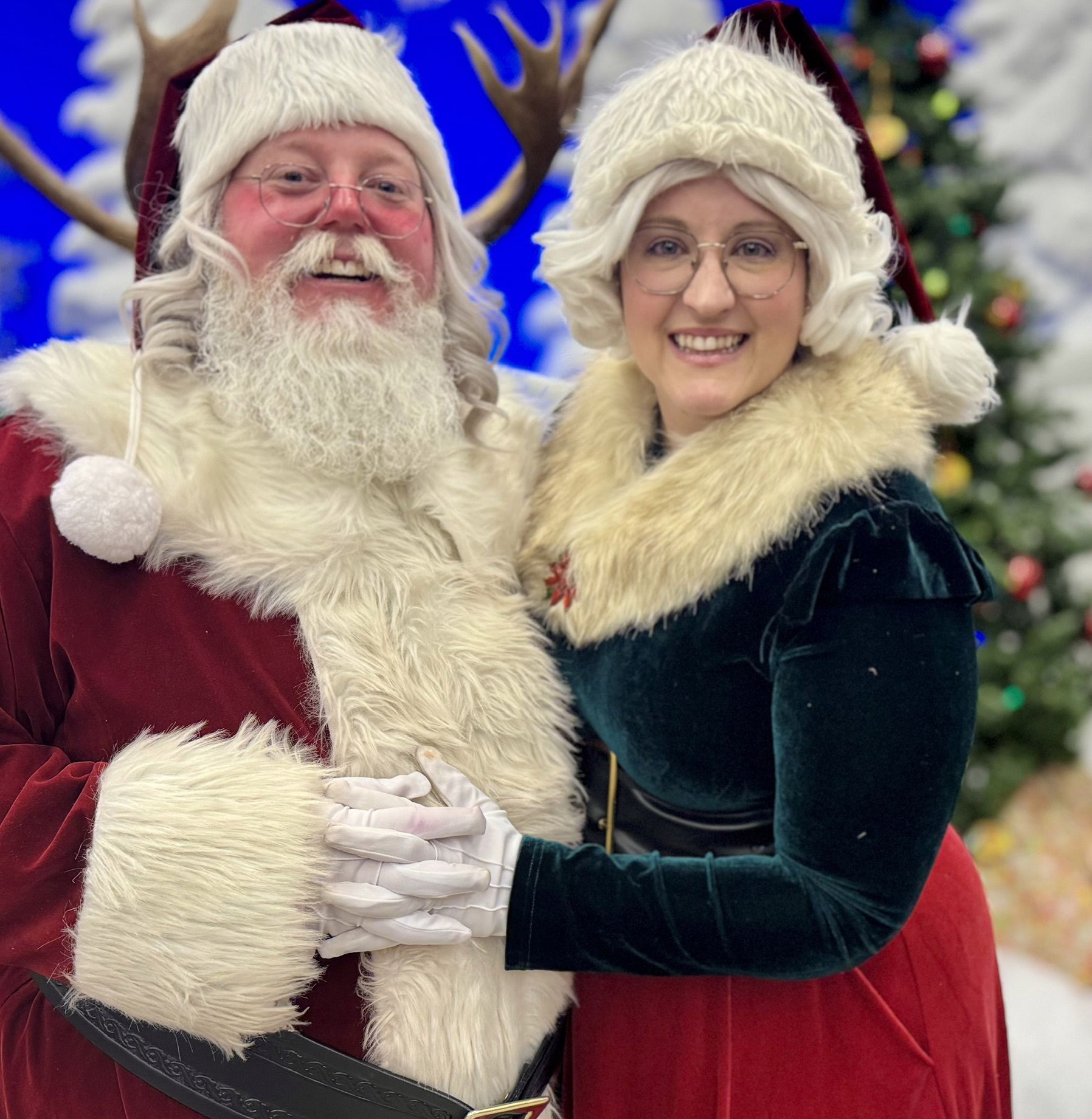 Santa Claus and a woman in festive attire smiling, with Christmas decorations in the background.