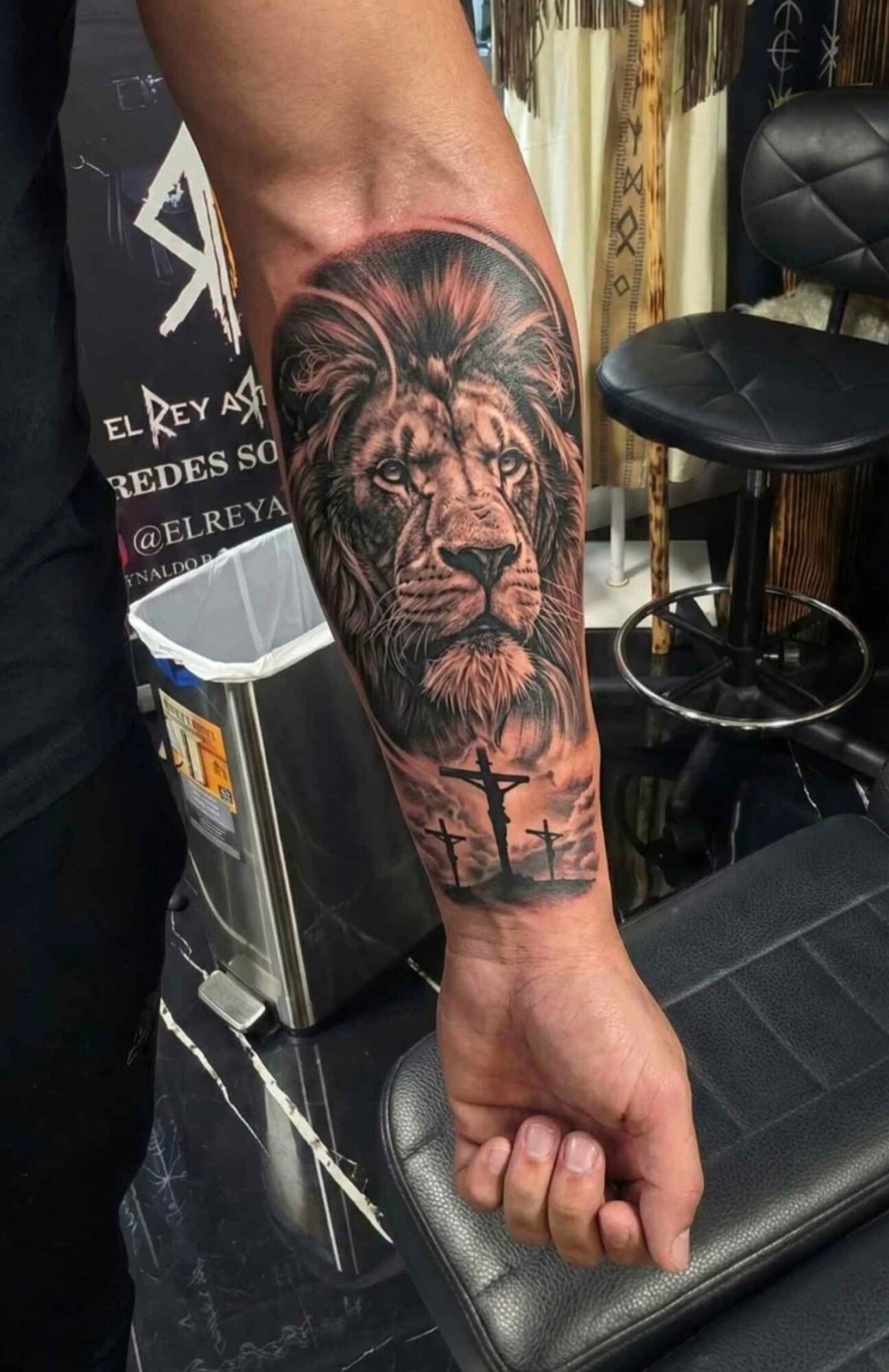 Tattoo of a lion's face with a landscape background on person's forearm, with trees and a sunset, in black and gray ink.