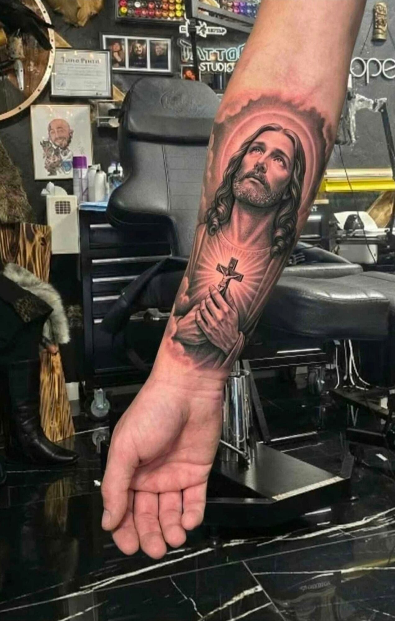 Tattoo of Jesus Christ with a halo, holding a cross, on a person's forearm, in a tattoo shop.