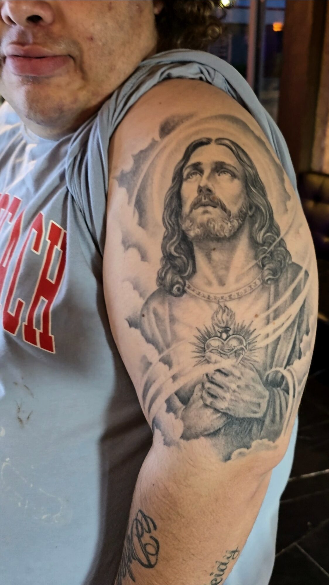 Tattoo of Jesus Christ with a radiant heart on upper arm, detailed face, long hair, and clouds background.