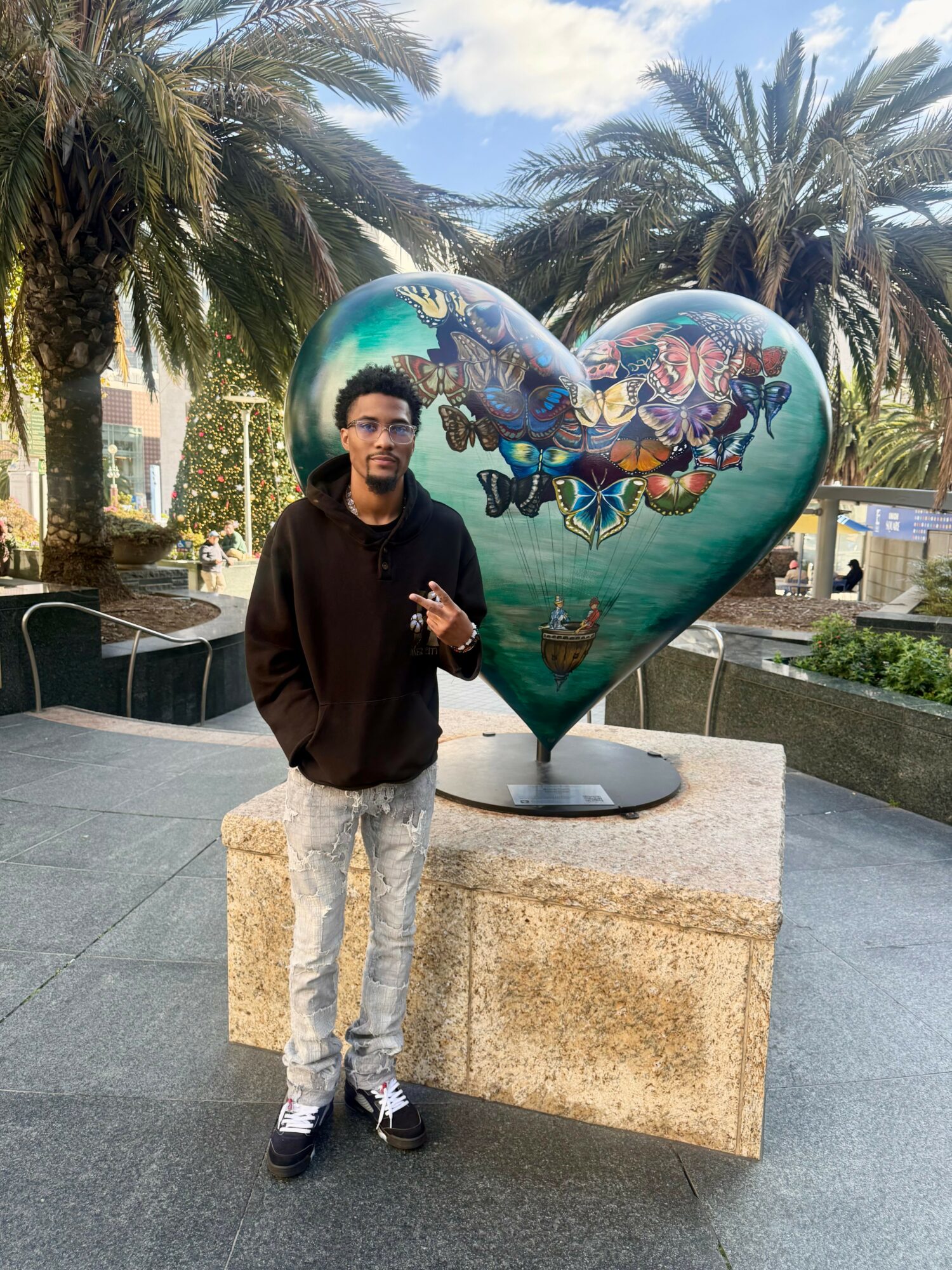 Young man standing outdoors in front of a large heart-shaped sculpture with colorful artwork, palm trees in background.