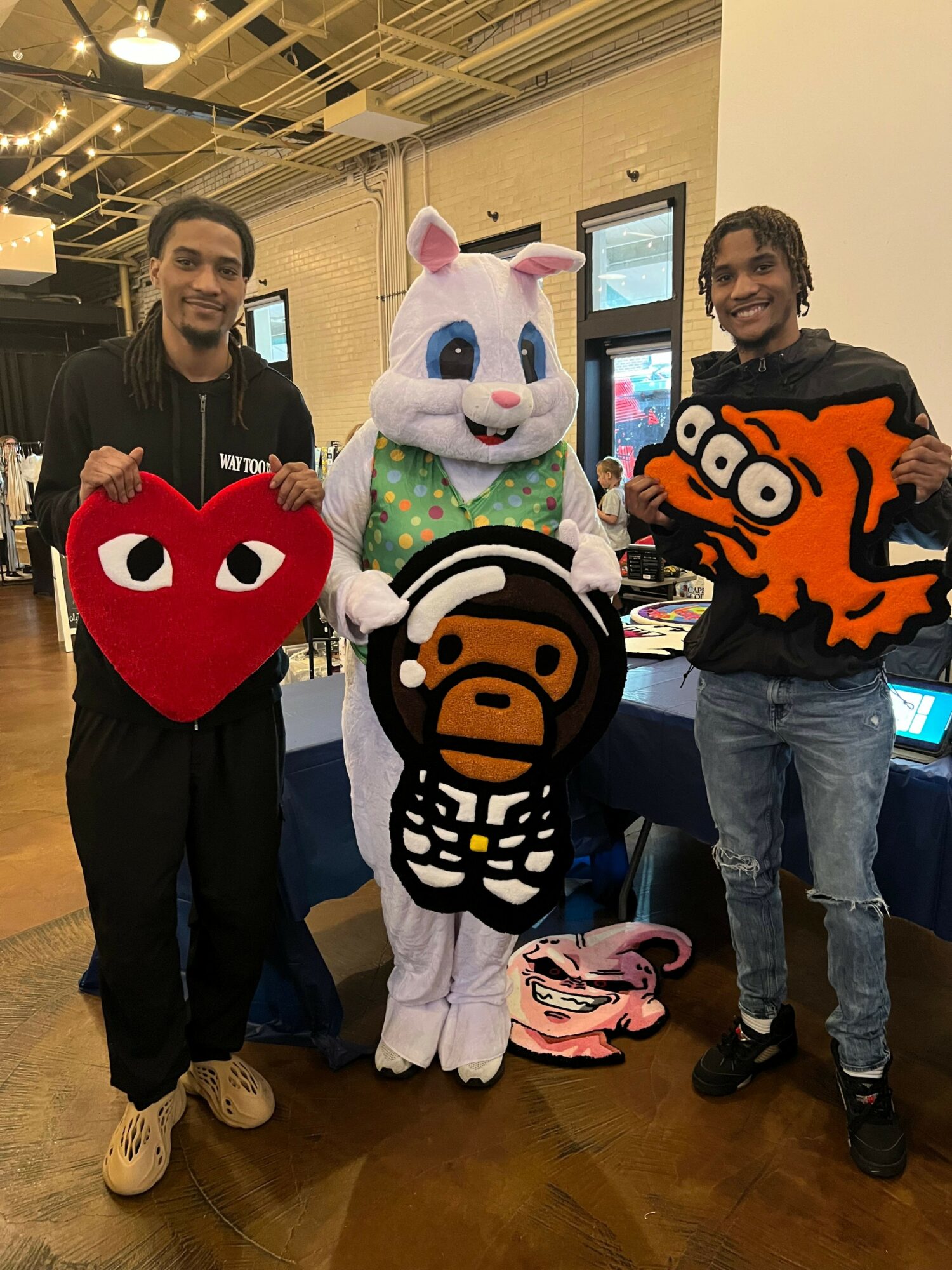 Two people in costumes, one with a unicorn head and another with a red heart-shaped head, standing indoors with computers and chairs.
