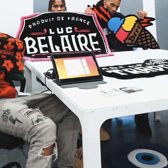 People sitting at a table with colorful patches and a tablet, with a sign reading 'LUC BELAIRE' behind them.