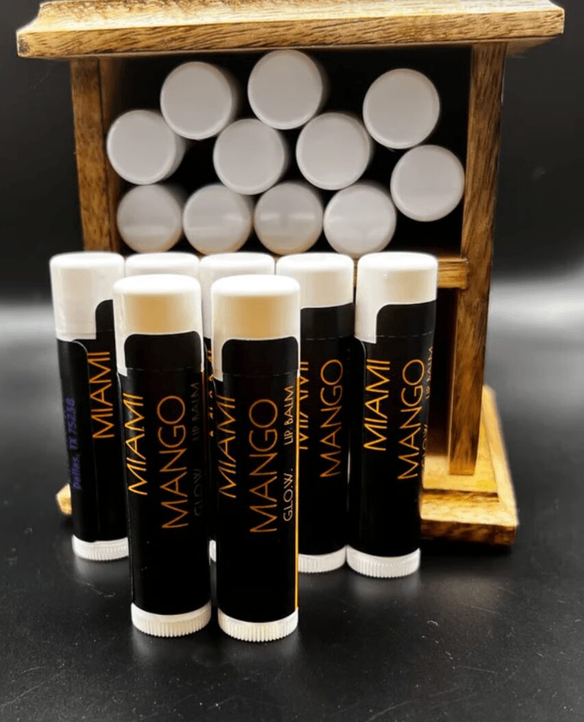 Eight lip balm sticks with black and gold labels in front of a wooden display holding more lip balms, all with white caps.