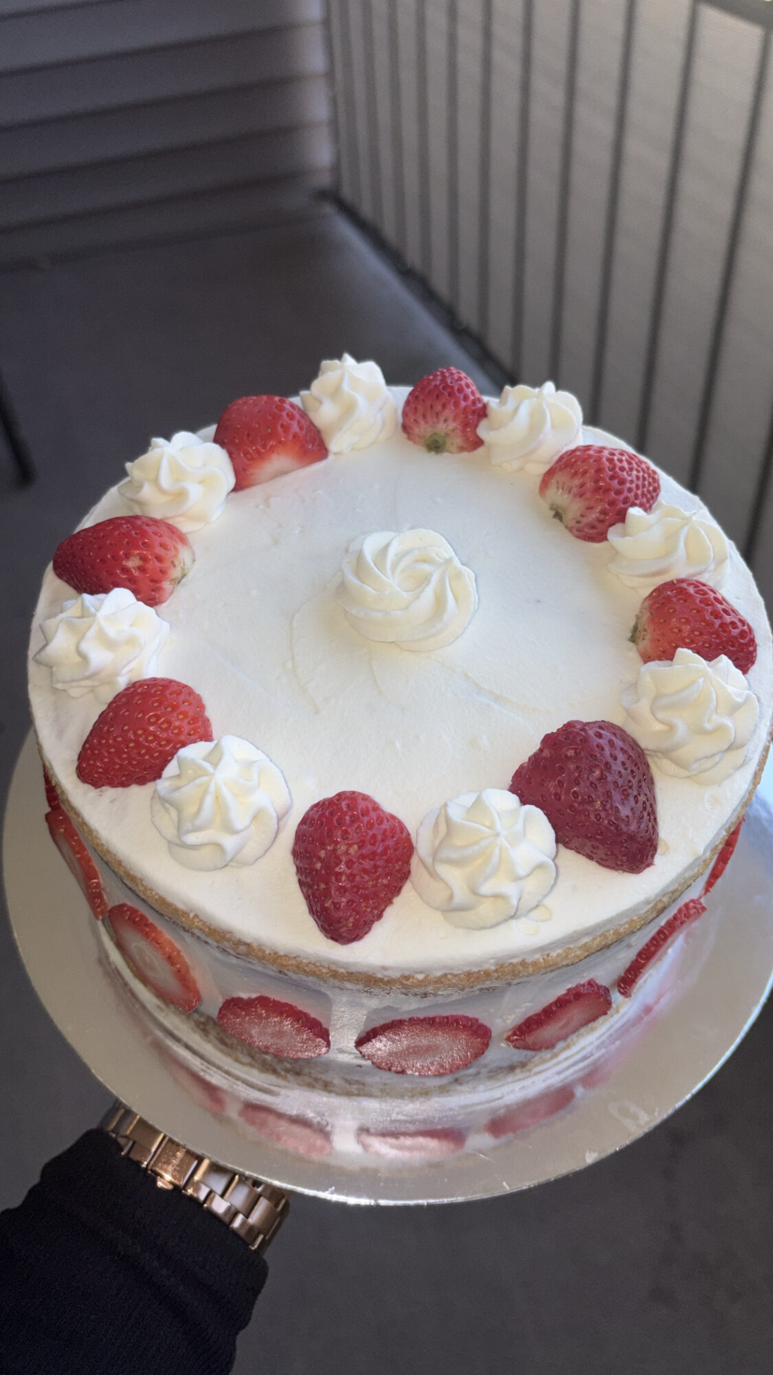 Round cake with white frosting, strawberries, and whipped cream decorations, held by a person in a dark sleeve, background of a gray wall.