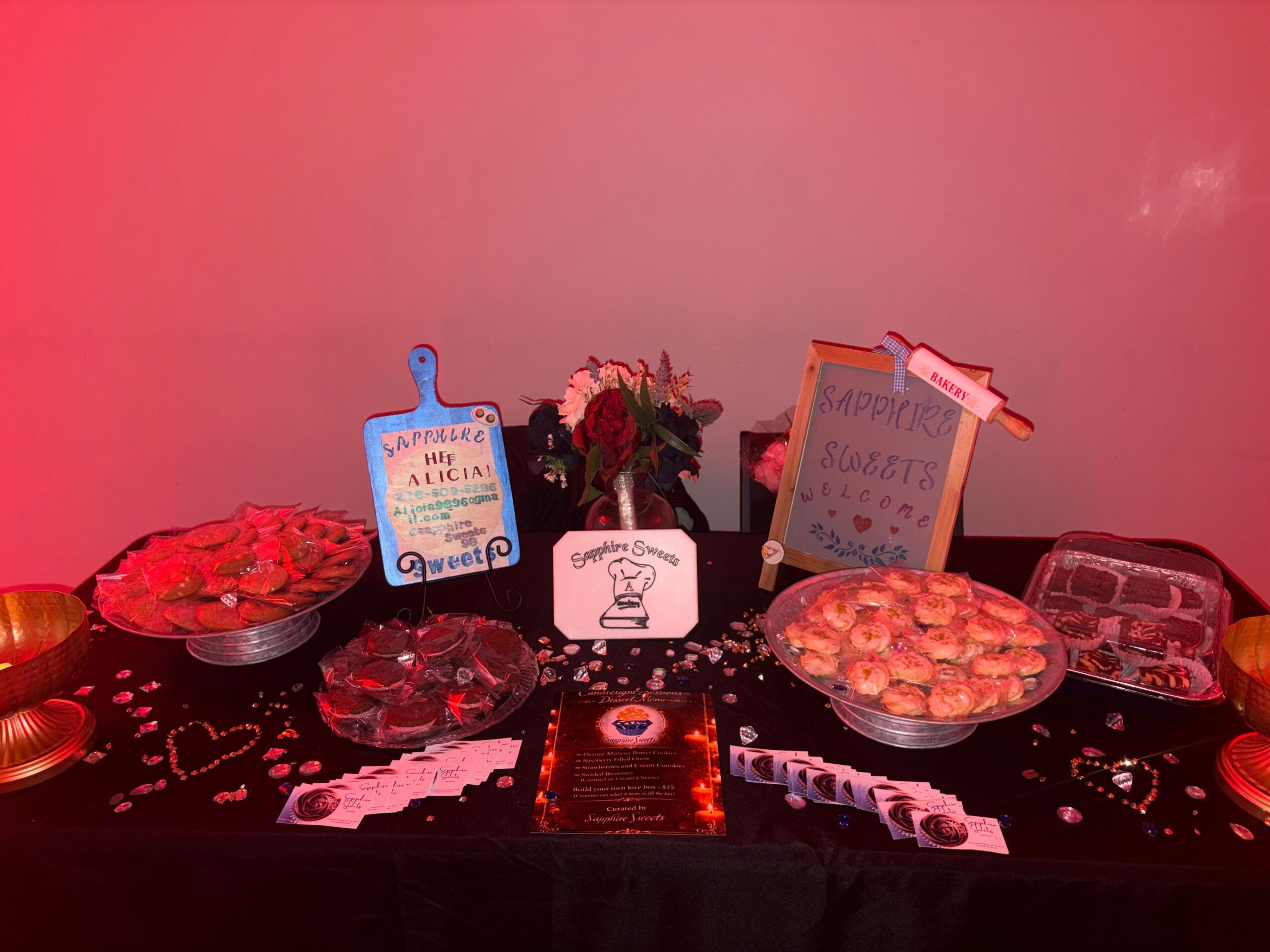 Table with flowers, signs, and snacks for celebration, decorated with confetti and party items, against a pink background.