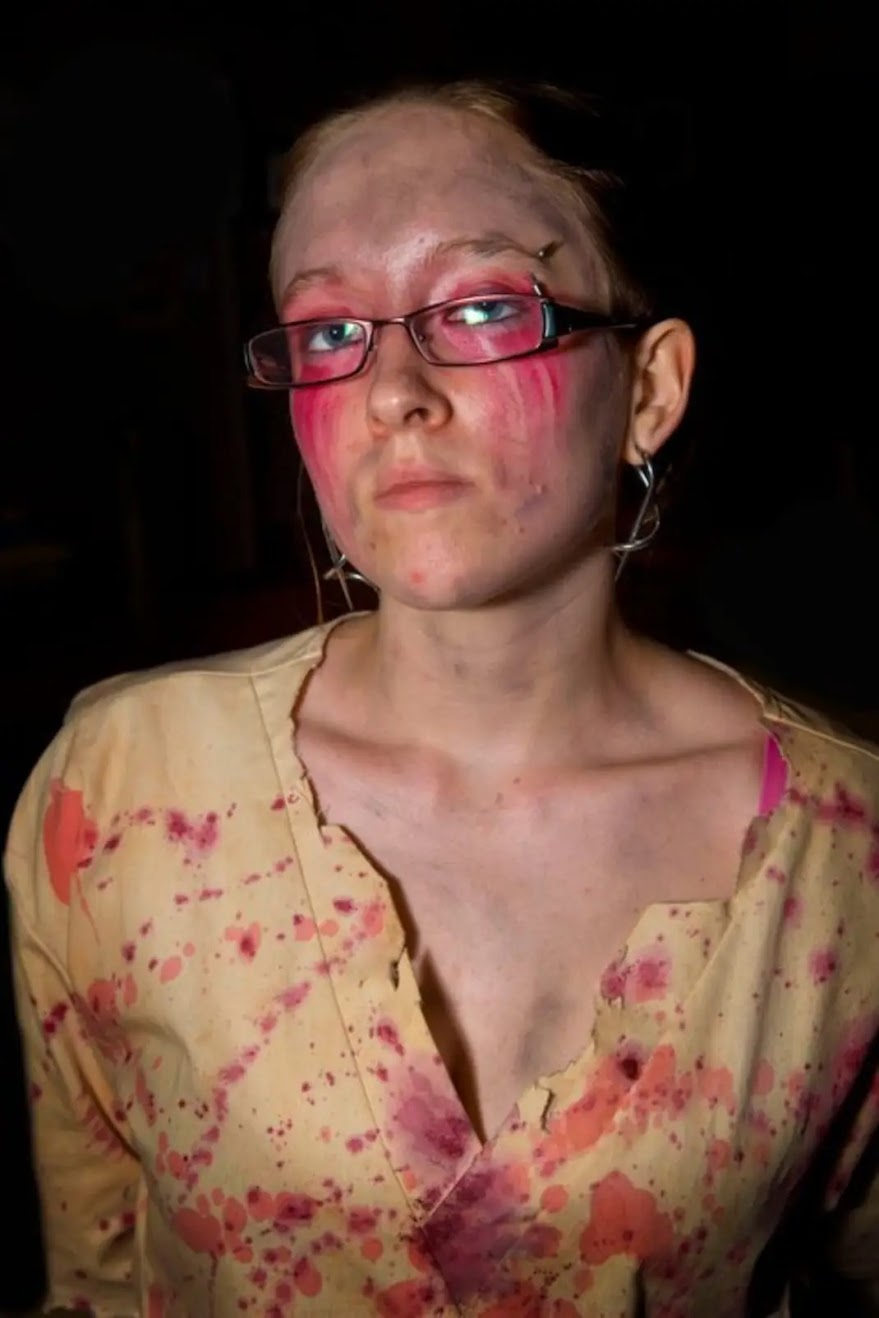 Young woman with glasses and makeup, wearing a yellow shirt with pink stains, looking serious against a dark background.