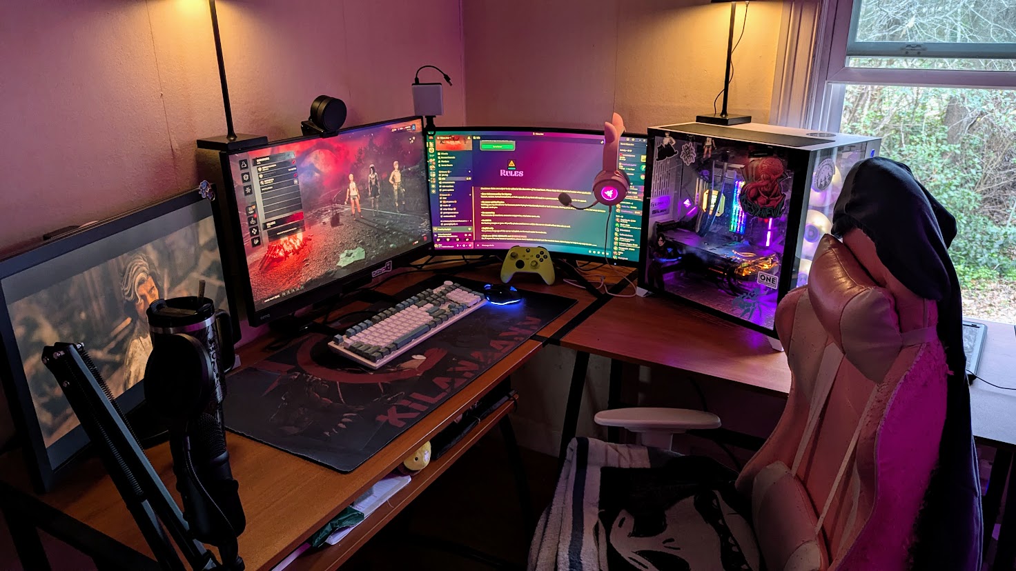 Computer desk with three monitors, keyboard, mouse, and gaming controller, in a room with a window and a chair, illuminated by warm lighting.