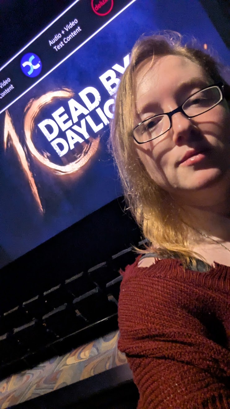 Young woman with glasses and long hair taking a selfie in front of a screen displaying 'DEAD BY DAYLIGHT'.