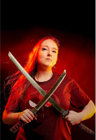 Woman with long red hair holding two swords crossed in front of her chest, dark background, intense expression.