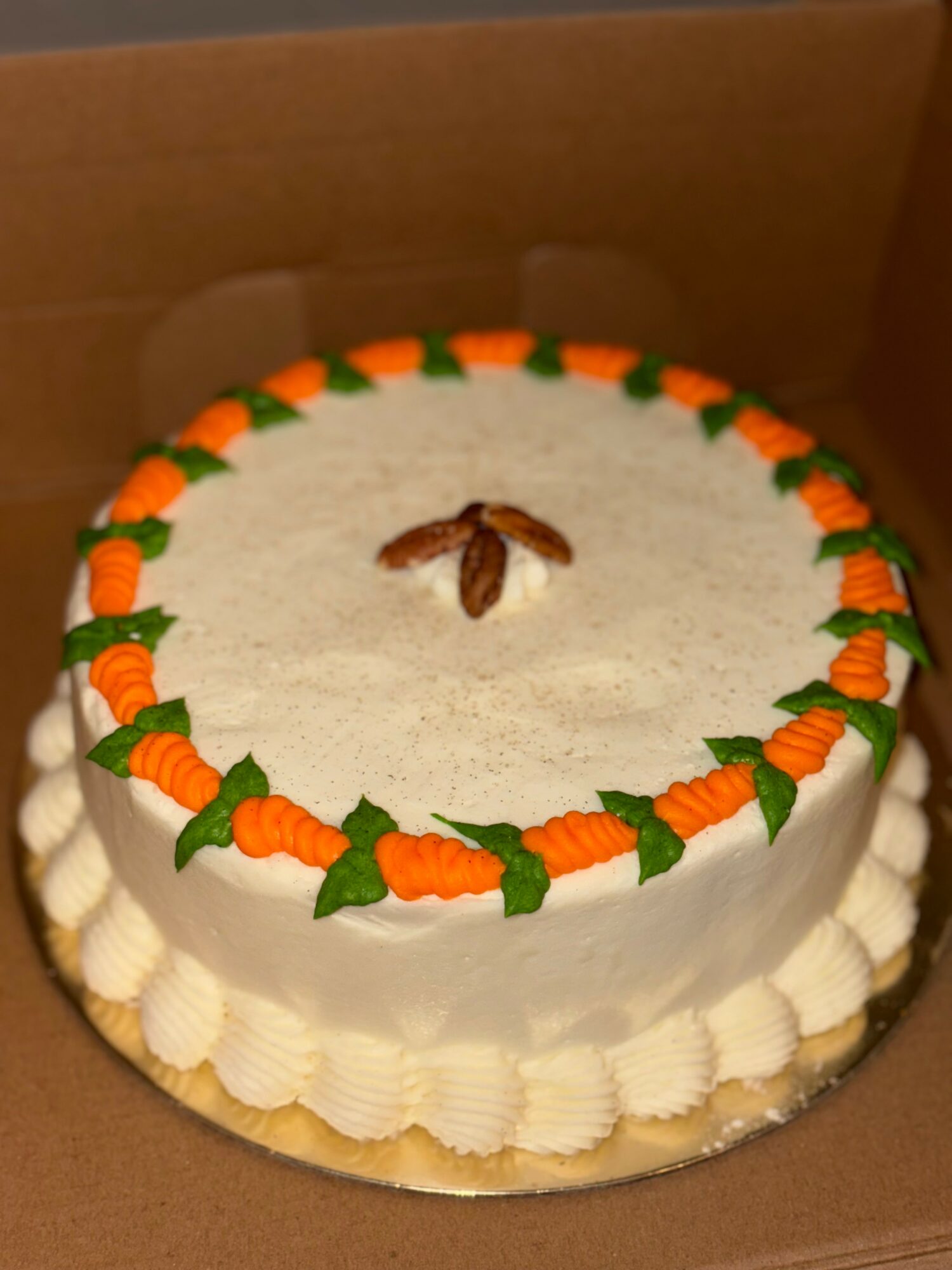 Round cake with white frosting, decorated with orange carrots and green leaves around the top edge, and a small brown decoration in the center.