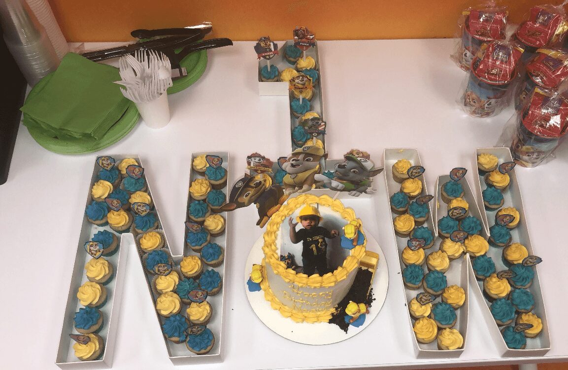 Celebration table with a cake, decorated letters, and party favors, including a green container and wrapped candies, on a white surface.