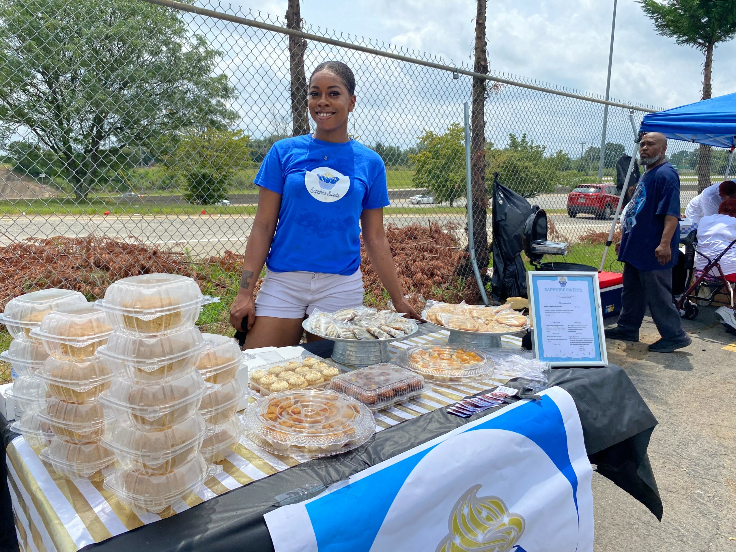 Smiling woman standing behind a table with baked goods at an outdoor market, with a chain-link fence and trees in background.