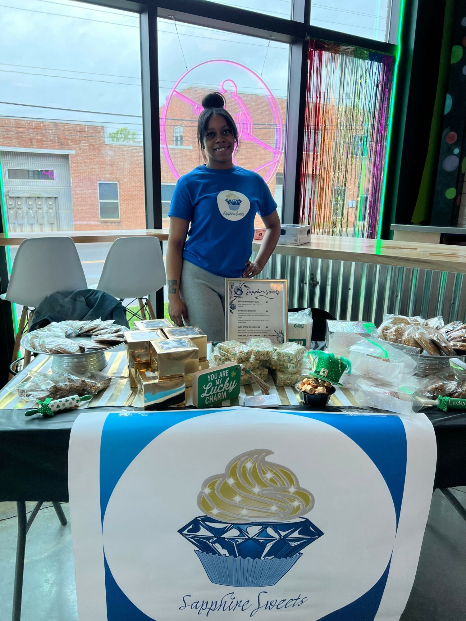 Person standing behind a table with various items, a large logo on a bag, and a window with colorful blinds.