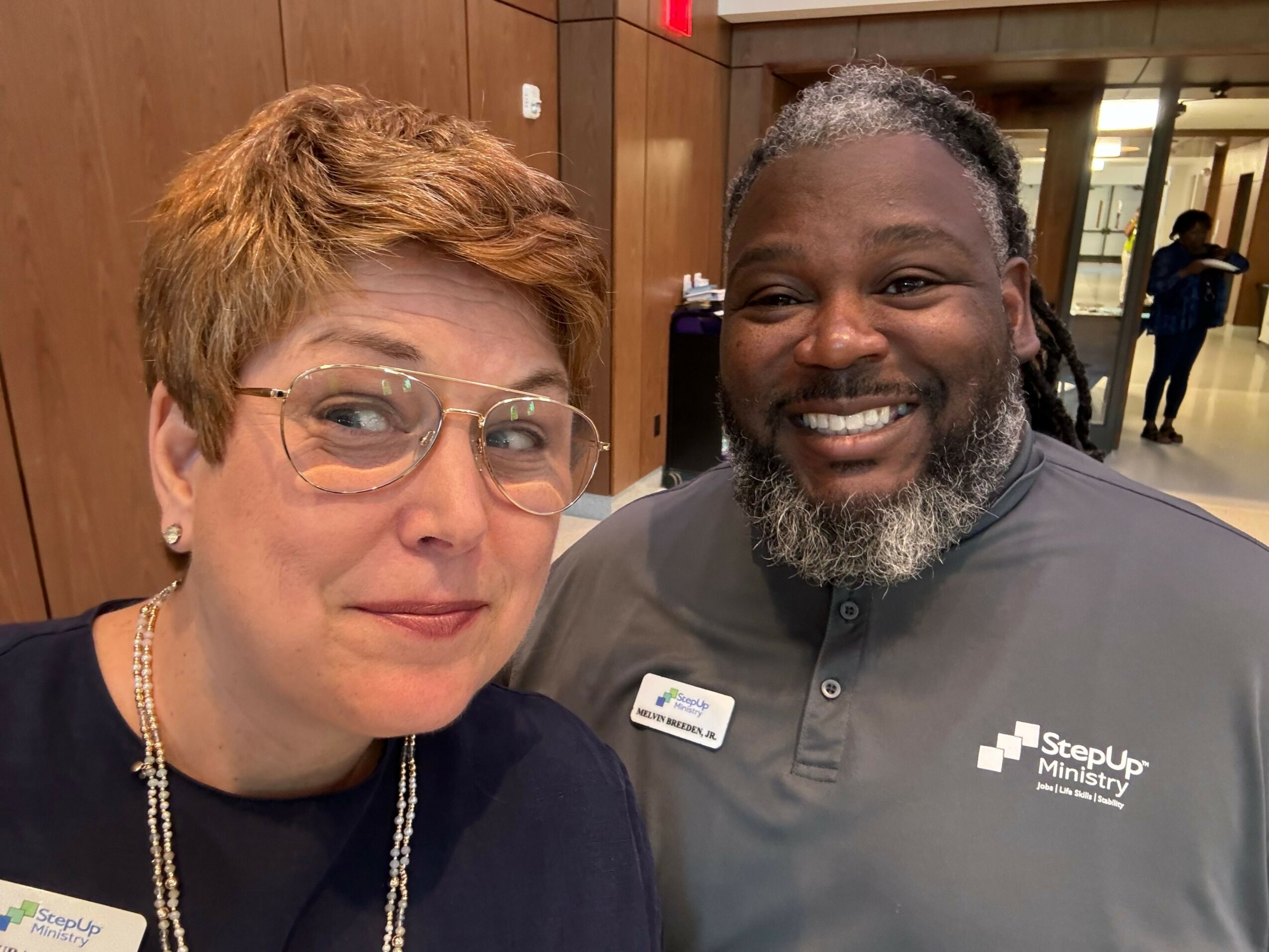 Two women smiling, one with glasses and short hair, the other with curly hair and a beard, in an indoor setting.
