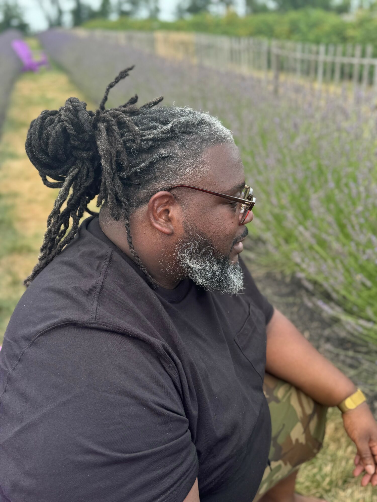 Profile of a man with dreadlocks, glasses, and a beard, sitting outdoors with a lavender field in background.