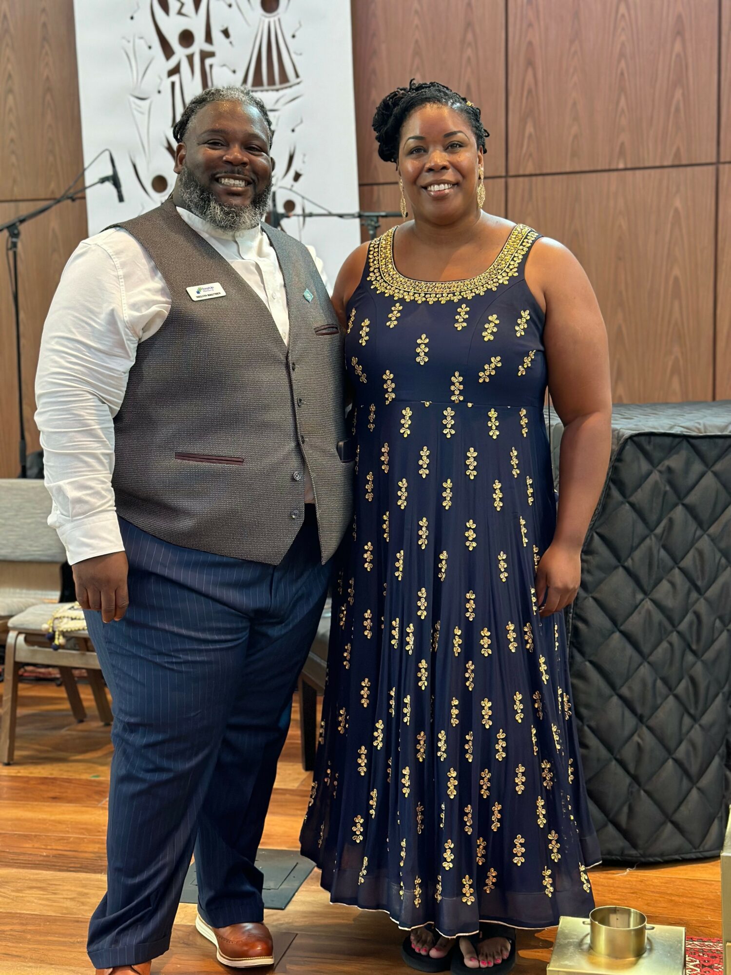 A man and woman stand together indoors, smiling, with a wooden wall and decorative elements behind them.