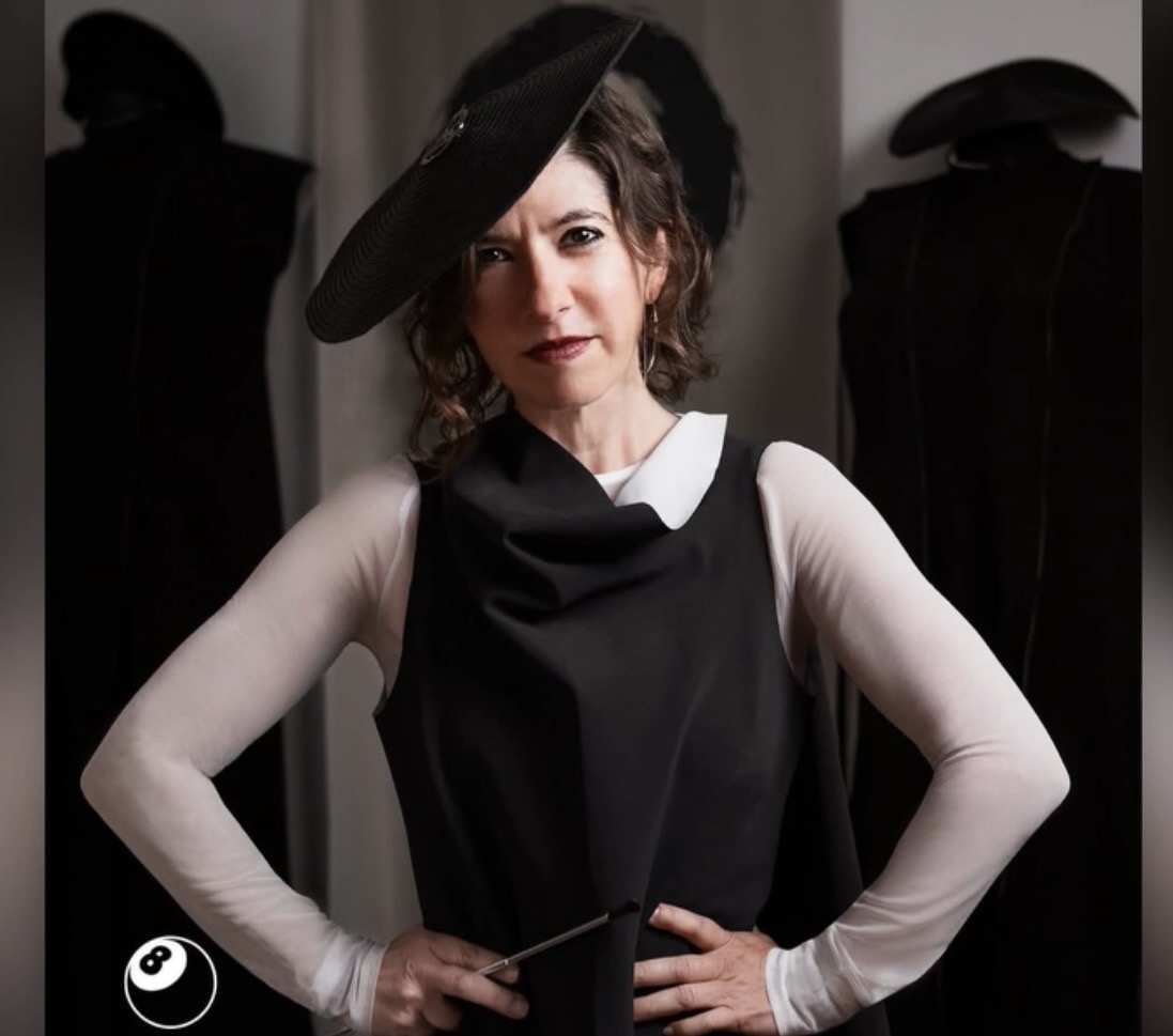 Woman wearing a black hat and dress, standing with hands on hips, in front of dark background.