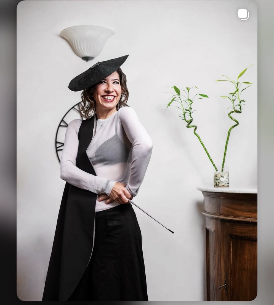 Woman smiling, wearing a hat, standing next to a wooden cabinet with a plant, clock, and wall light behind her.