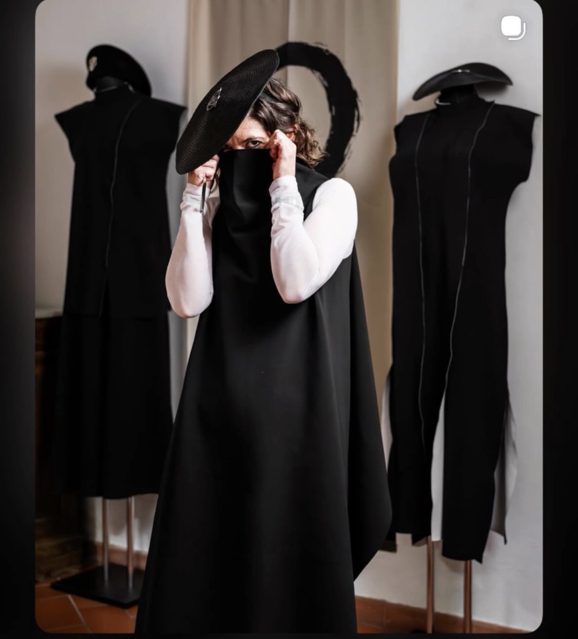 Person wearing a black hat and dress, adjusting glasses, with two black mannequins dressed in black outfits behind them.