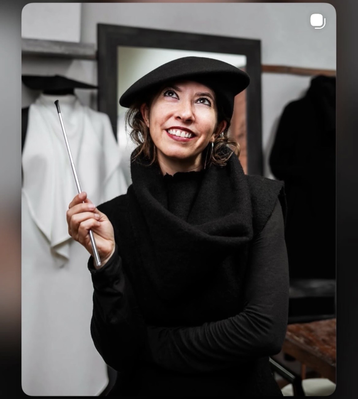 Woman smiling, wearing a hat and black coat, holding a shopping bag, in a clothing store, mirror in background.
