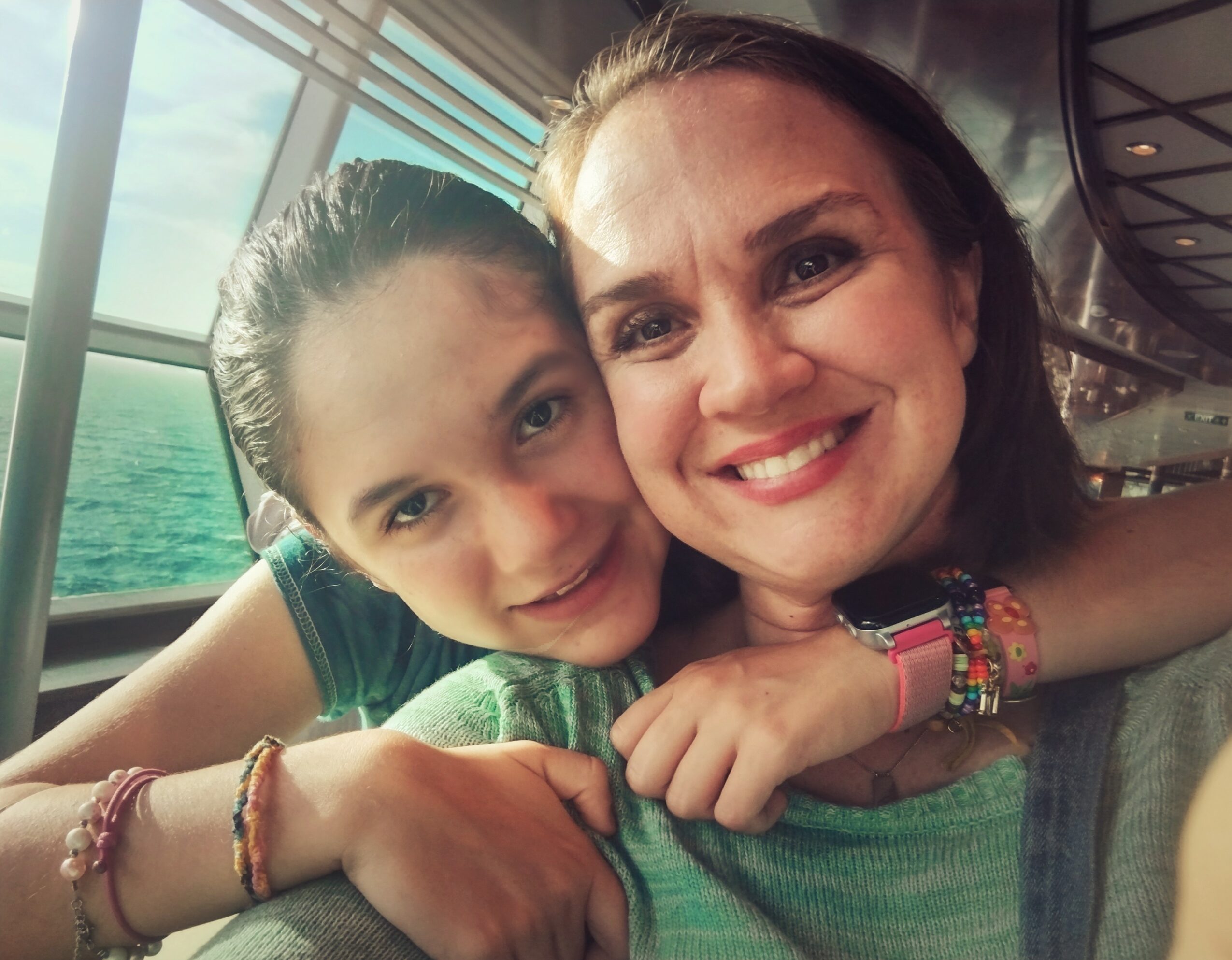 Two smiling females, one with dark hair and one with lighter hair, hugging inside a boat with water visible outside the window.