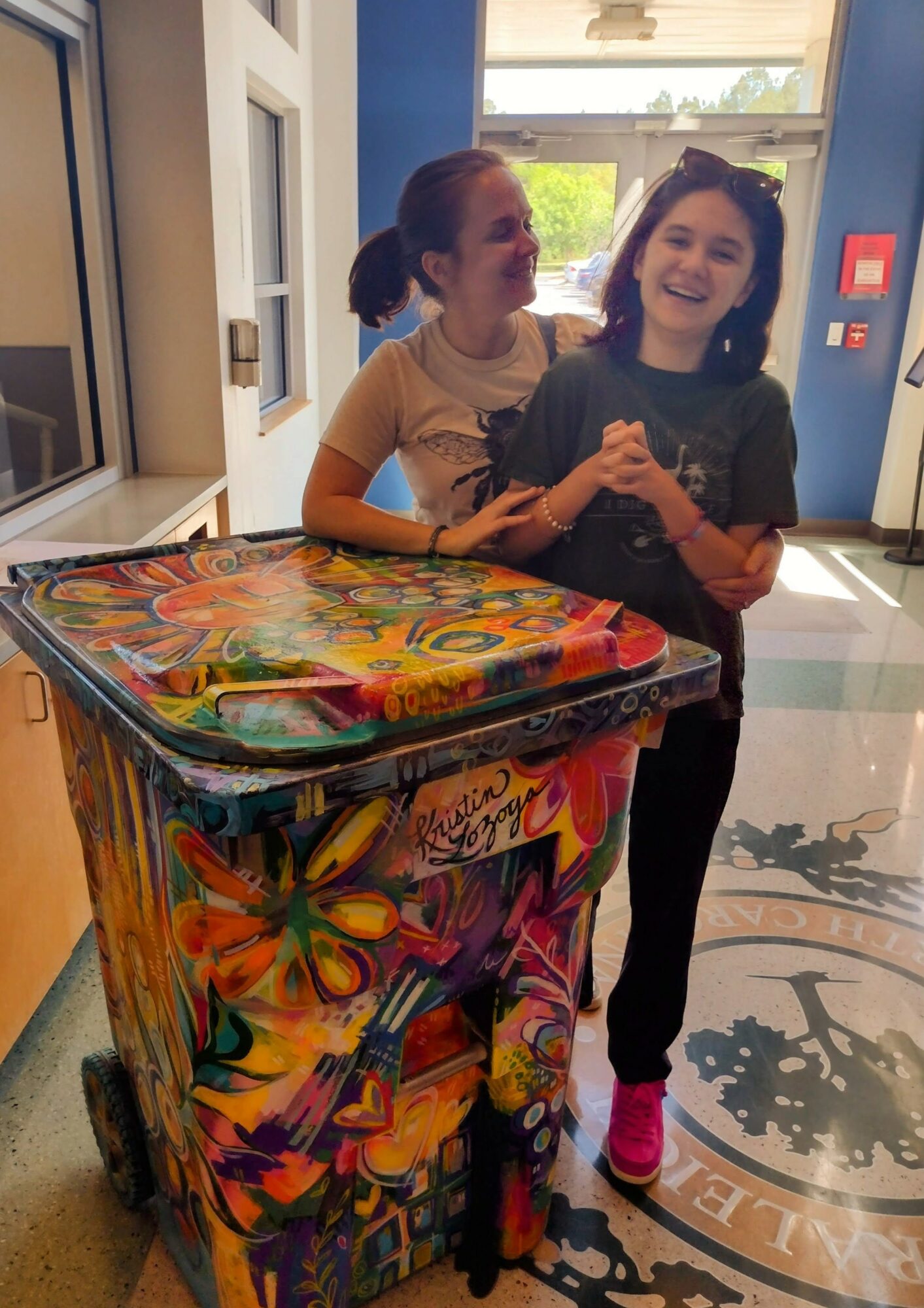 Two girls smiling and holding hands inside a building near a colorful cart, with a window and door in the background.