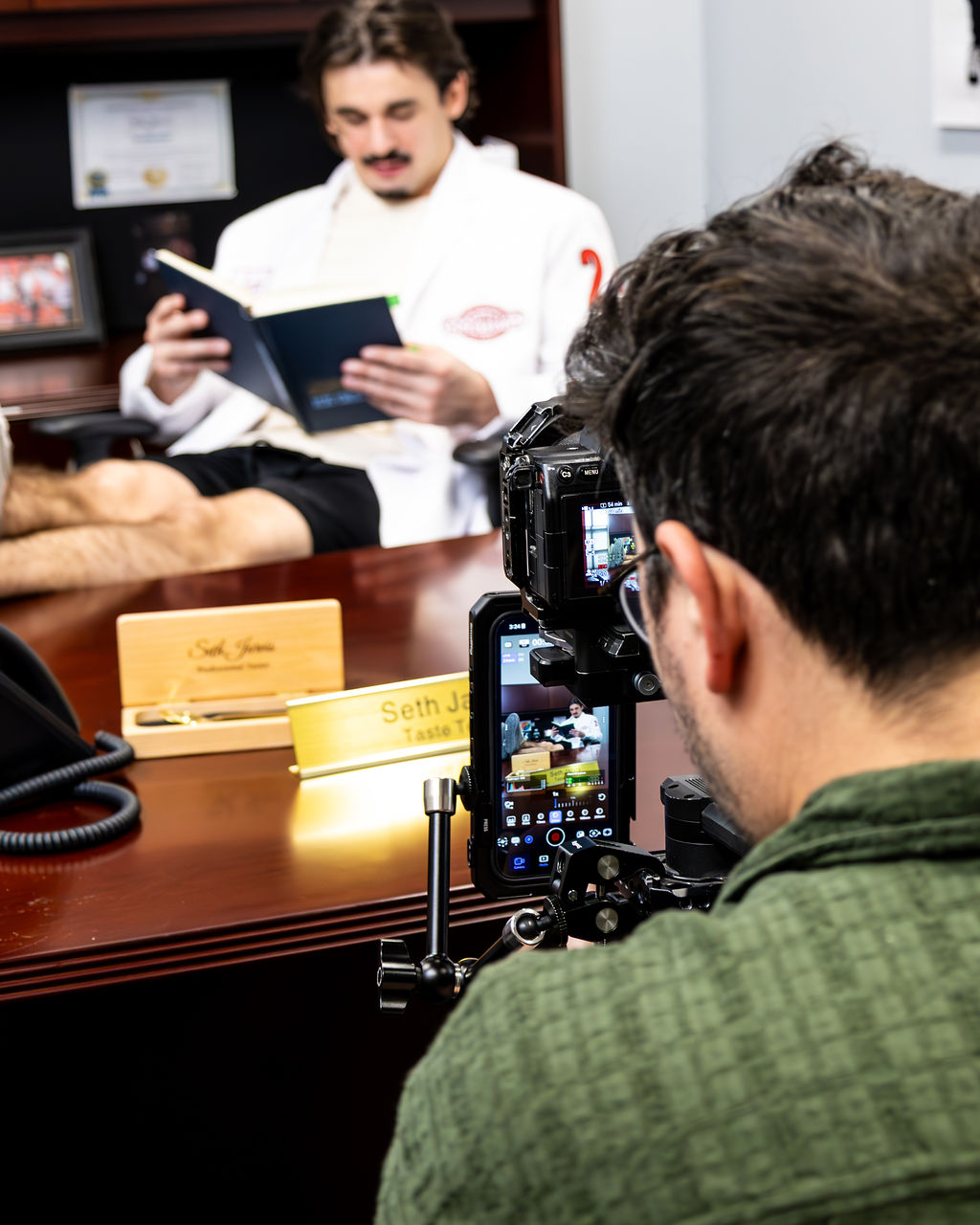 Doctor in white coat reading a book during an interview with a person taking photos, in an office setting.