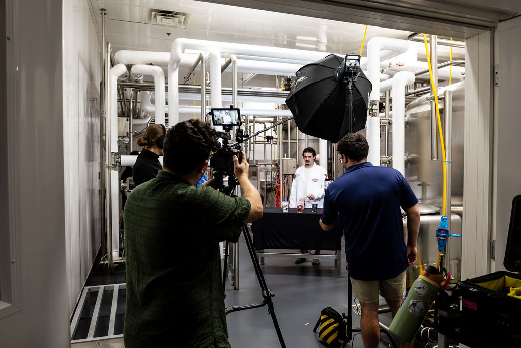 People filming a person in a room with pipes and equipment, with a large black umbrella light, in an industrial setting.