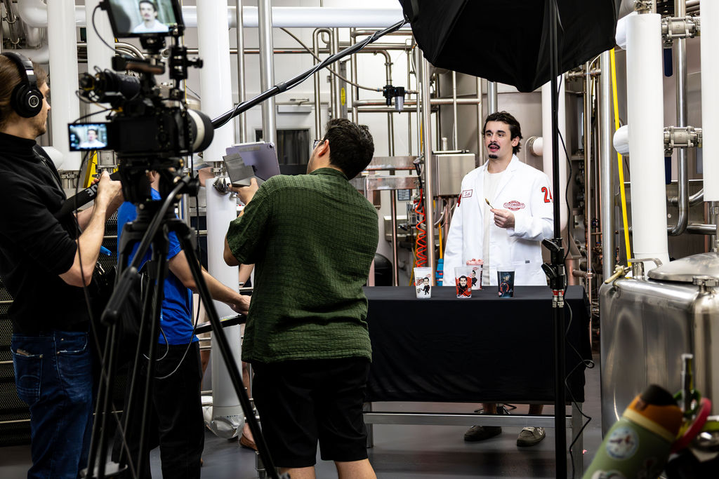 Person in a white lab coat demonstrates a science experiment to a group in a laboratory setting, with equipment and cameras present.