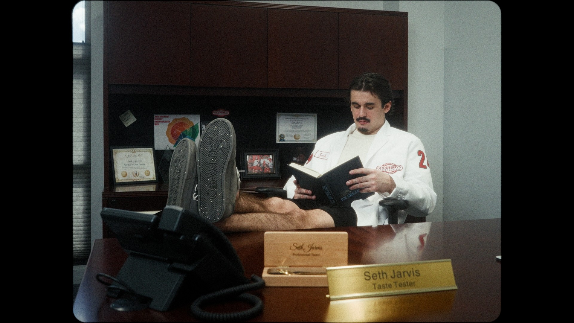 Person lying on desk with feet up, talking to a man in a white shirt holding a book, in an office setting.