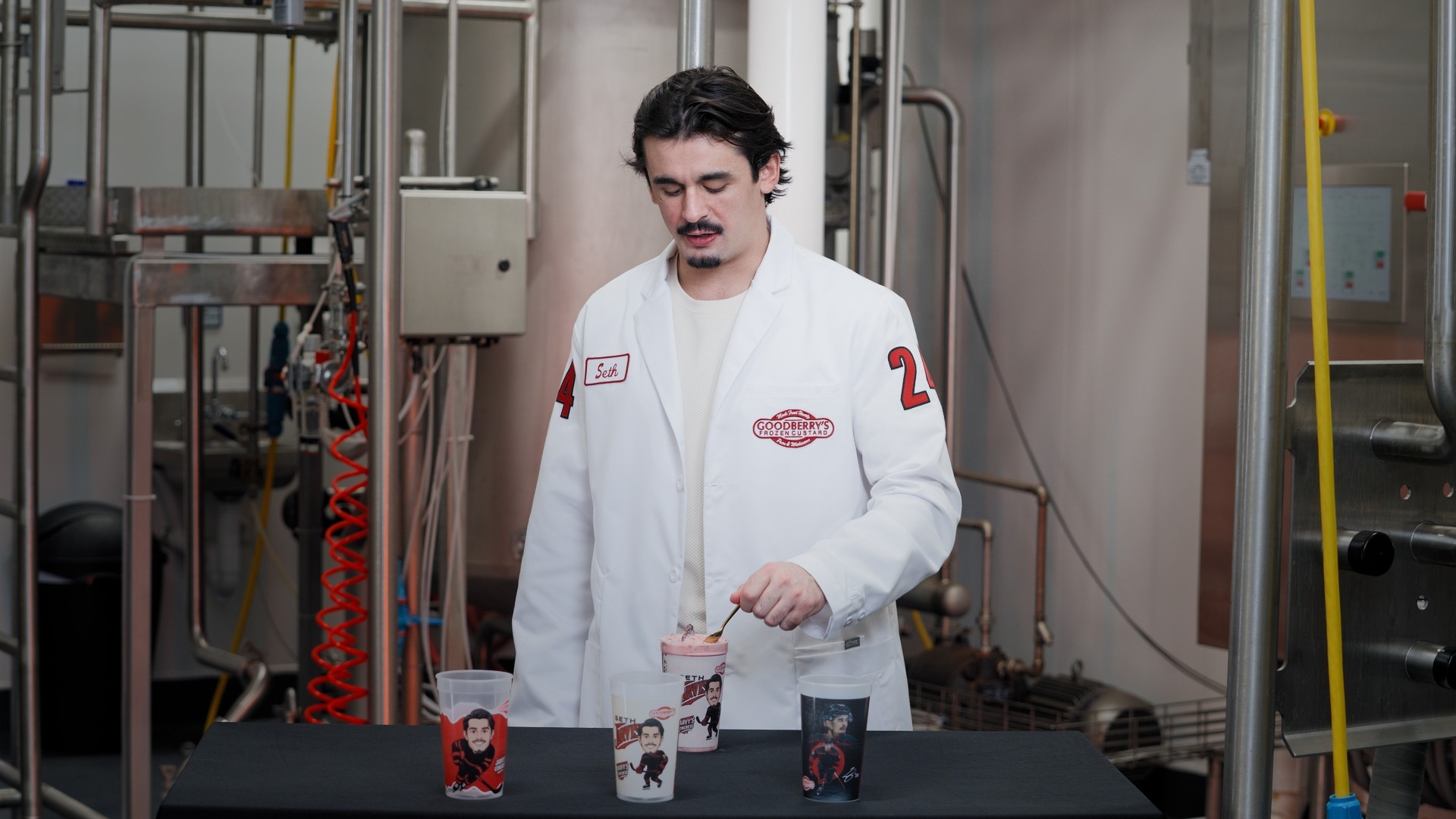 Man in a white lab coat standing behind a table with three small containers, in an industrial setting.