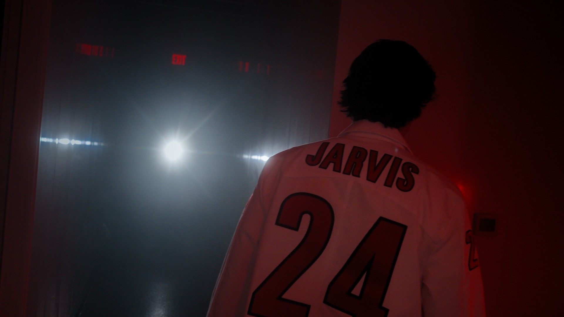 Person with curly hair wearing a sports jersey with the name 'JARVIS' and number 24, facing away in a dimly lit room.