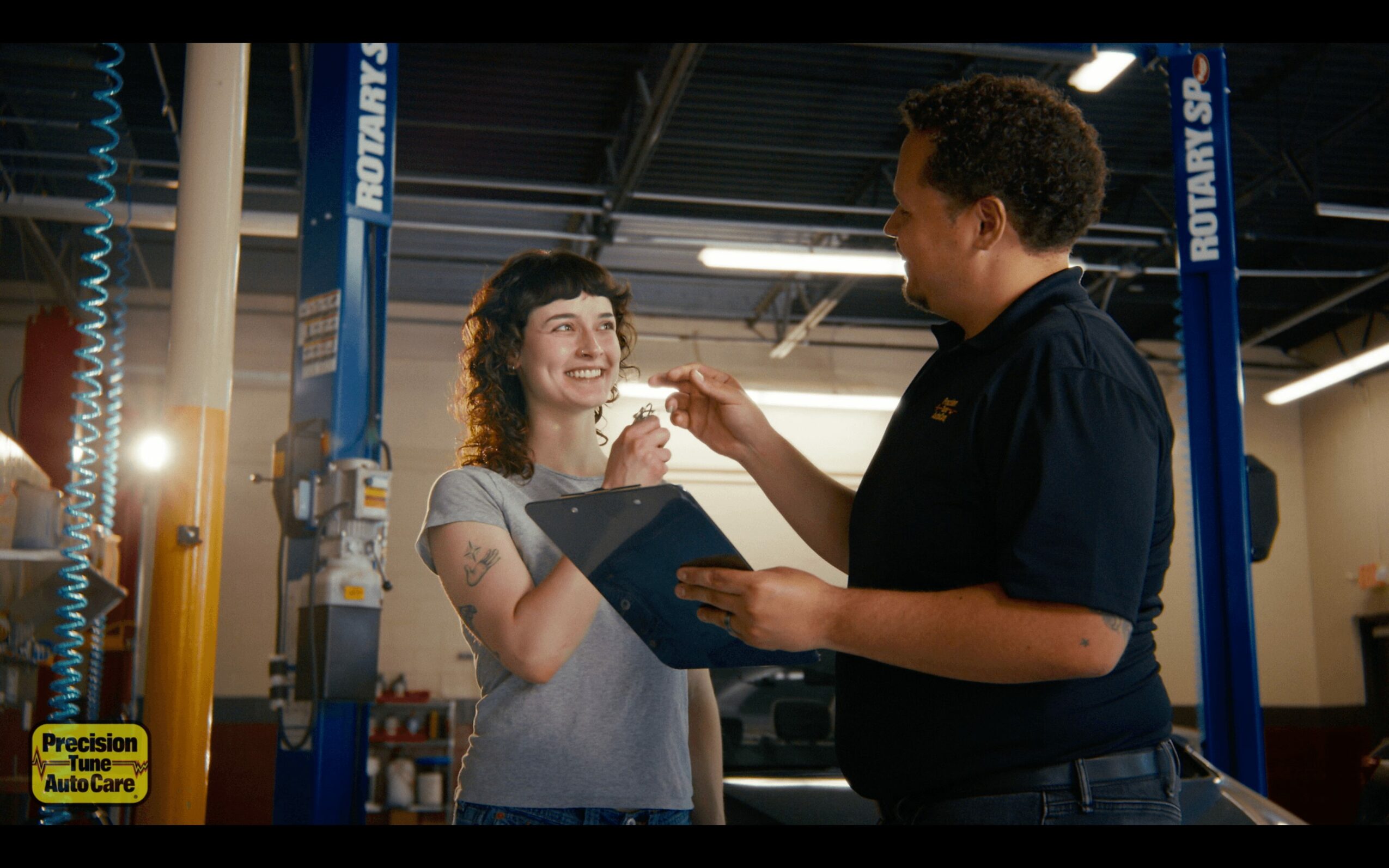 A woman and a mechanic are smiling and talking in a garage, with the mechanic holding a clipboard.