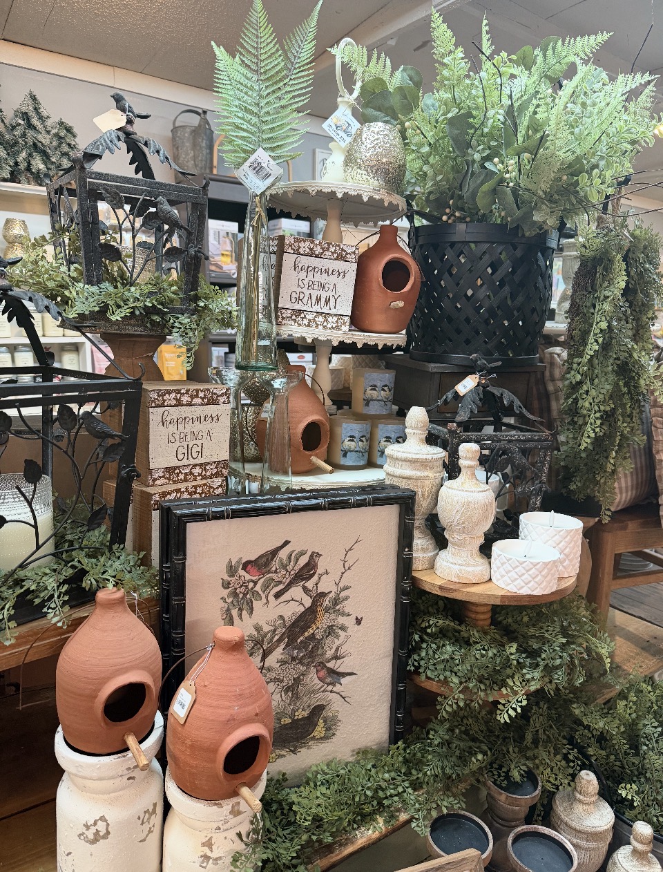 Display of decorative pottery, framed artwork, and lush green plants on shelves and tables in a store.