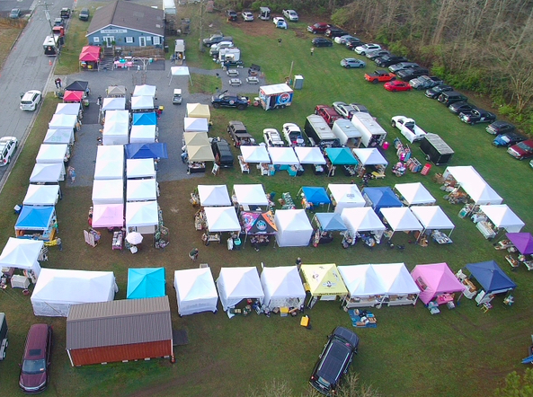Aerial view of an outdoor market with tents, cars, and a grassy area, viewed from the side. Word count: 20.