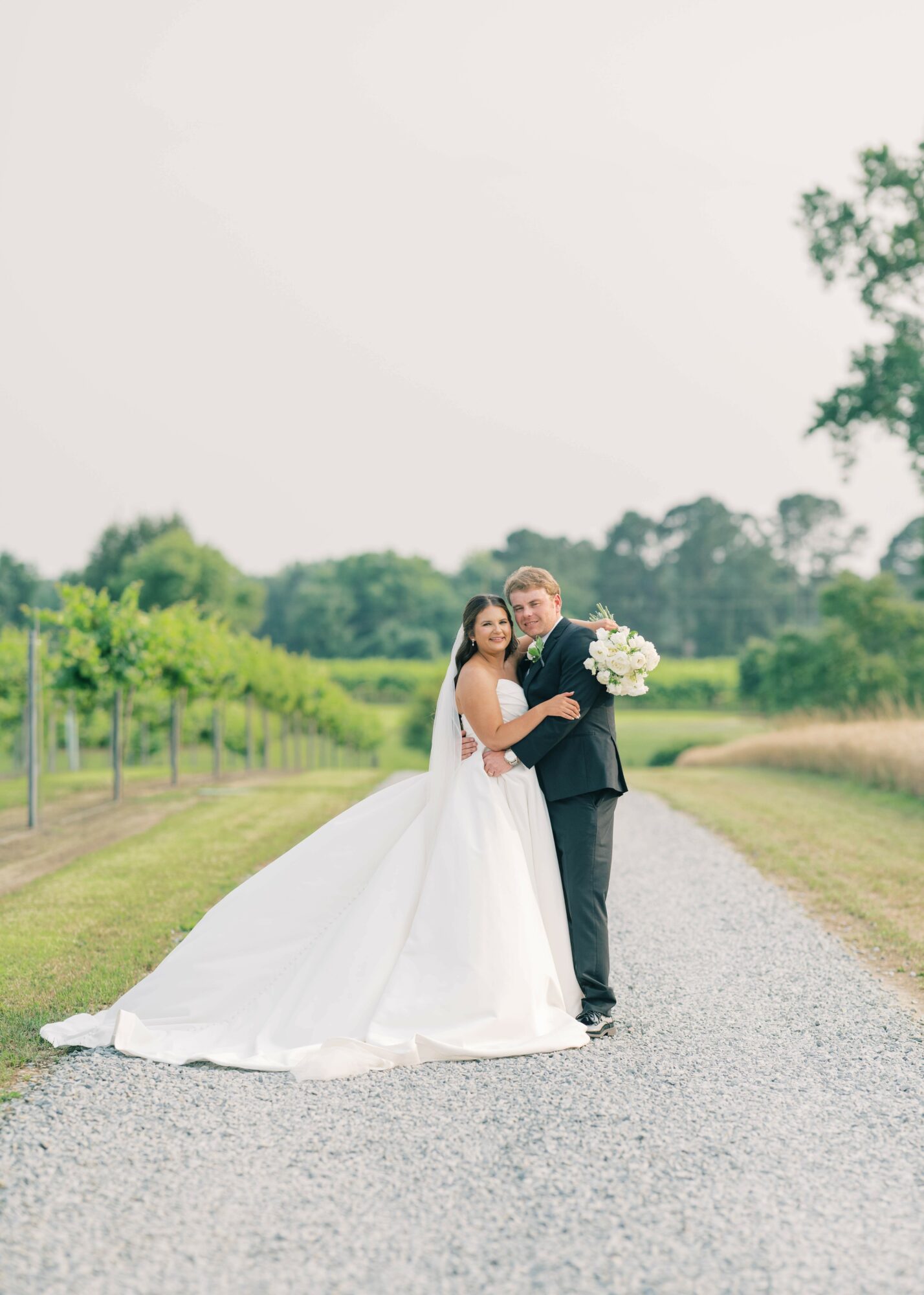 A bride and groom stand on a gravel path in a green outdoor setting, holding a bouquet of flowers, smiling.