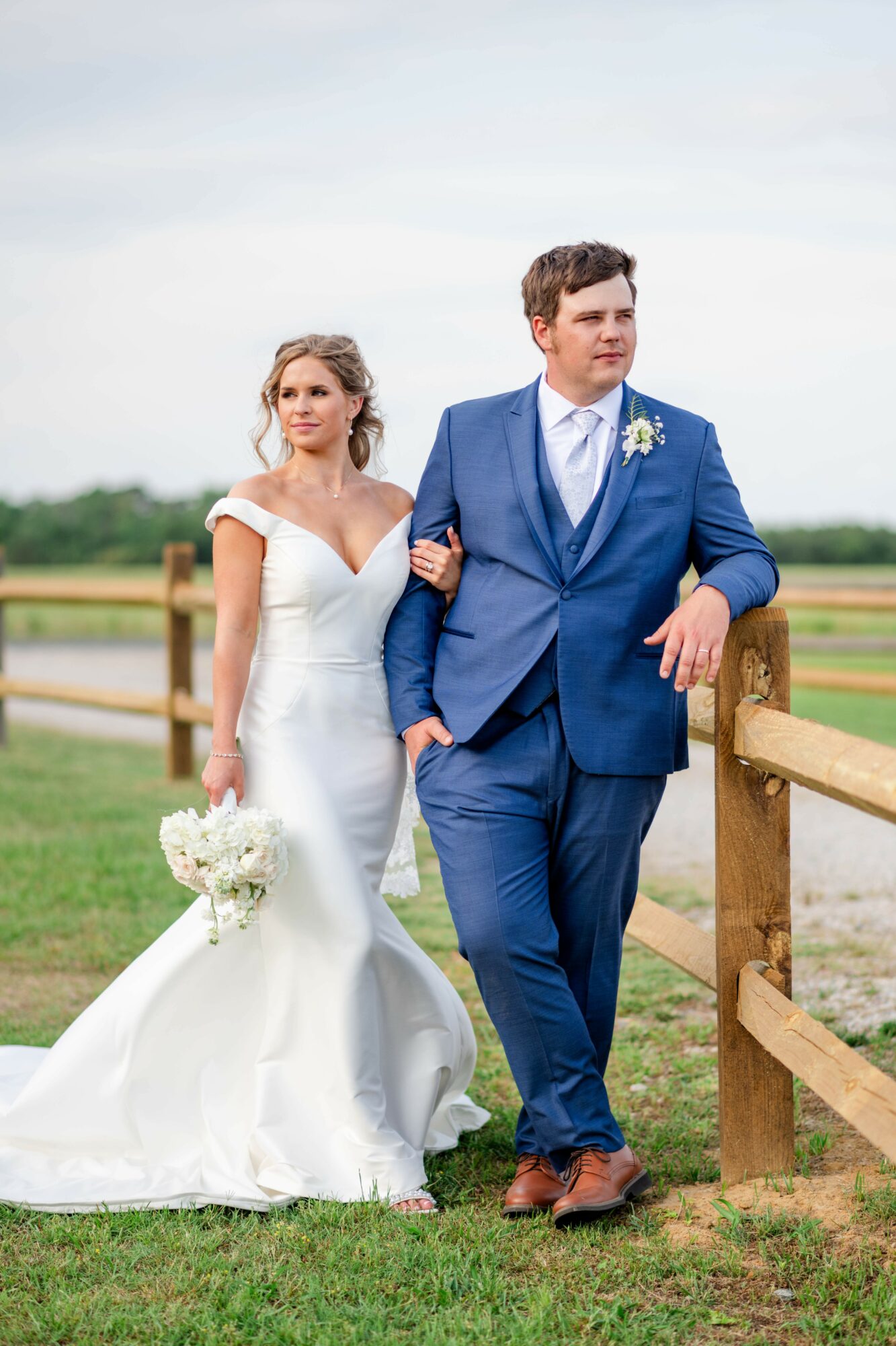 Bride in a white wedding dress holding a bouquet, groom in a blue suit walking outdoors near a wooden fence.