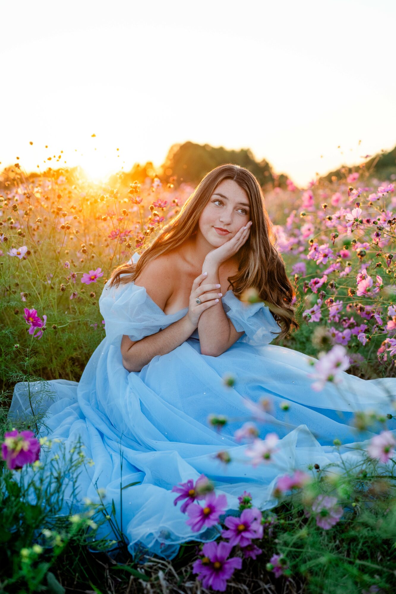 Young woman in a blue dress sitting in a flower field during sunset, resting her chin on her hand.