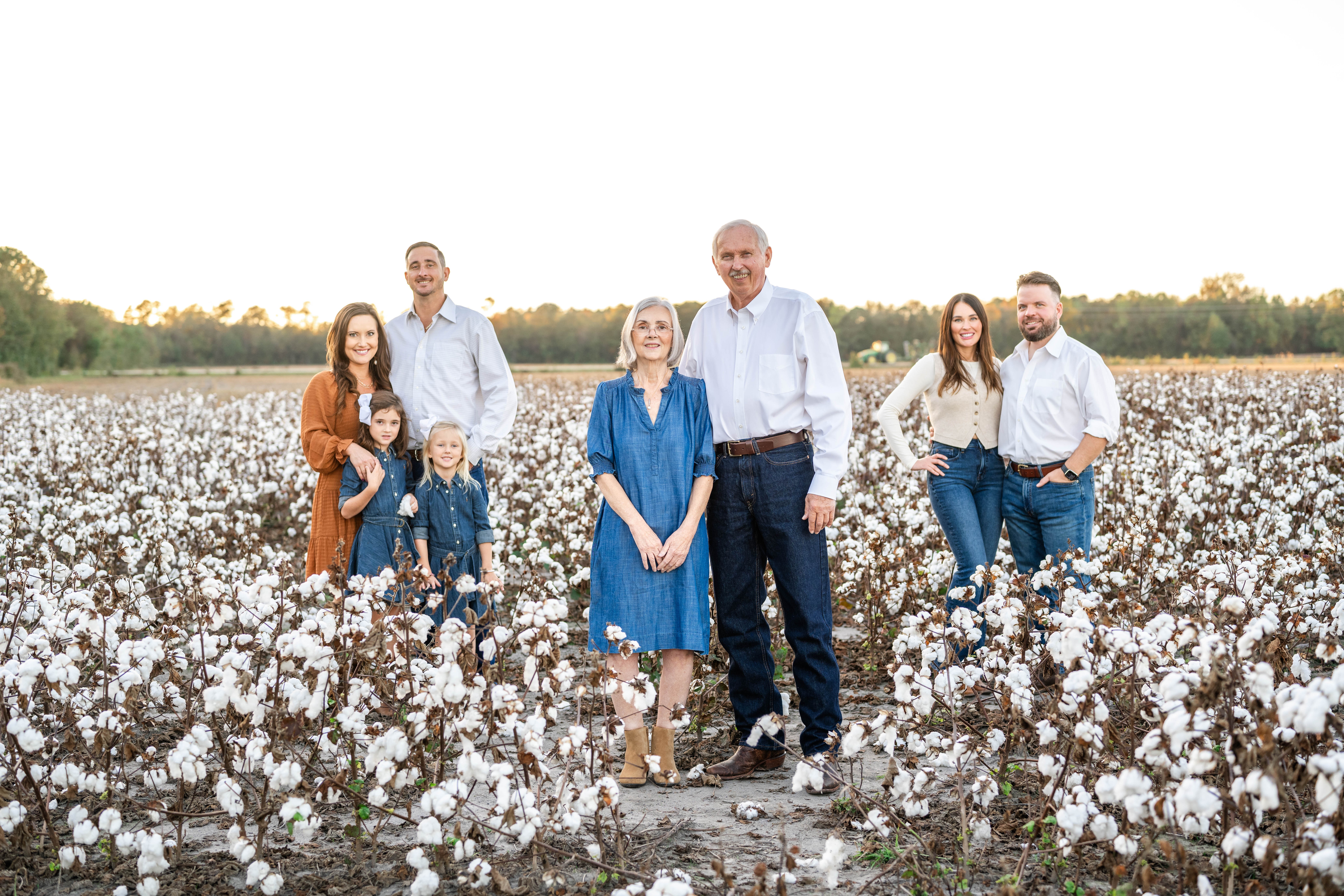 Group of seven people standing in a cotton field with trees in the background, smiling, during daytime.