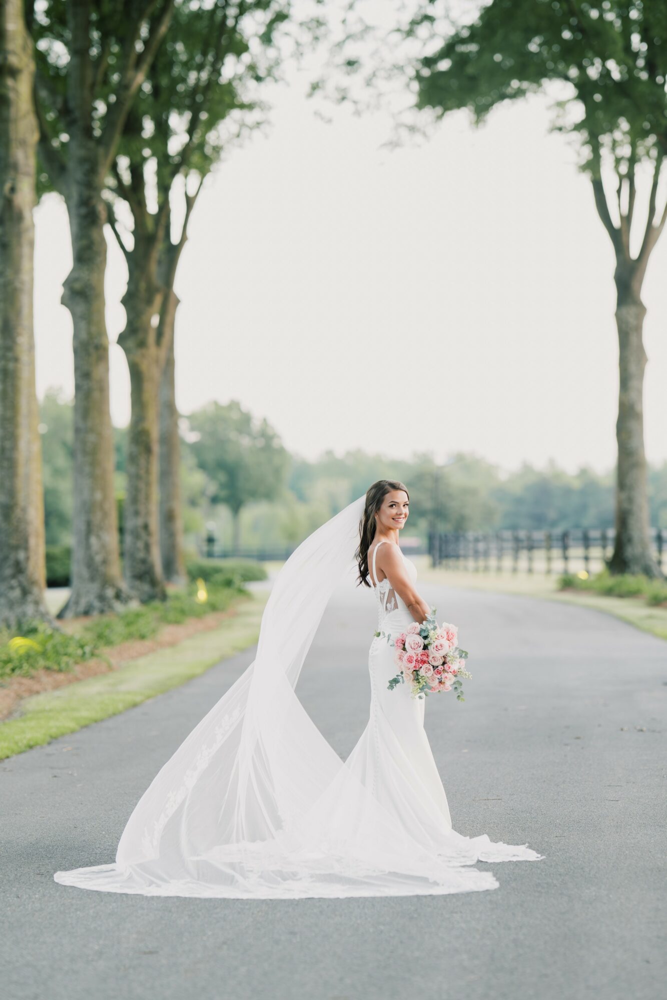 Bride in a white wedding dress holding a bouquet, standing on a road with trees in the background.