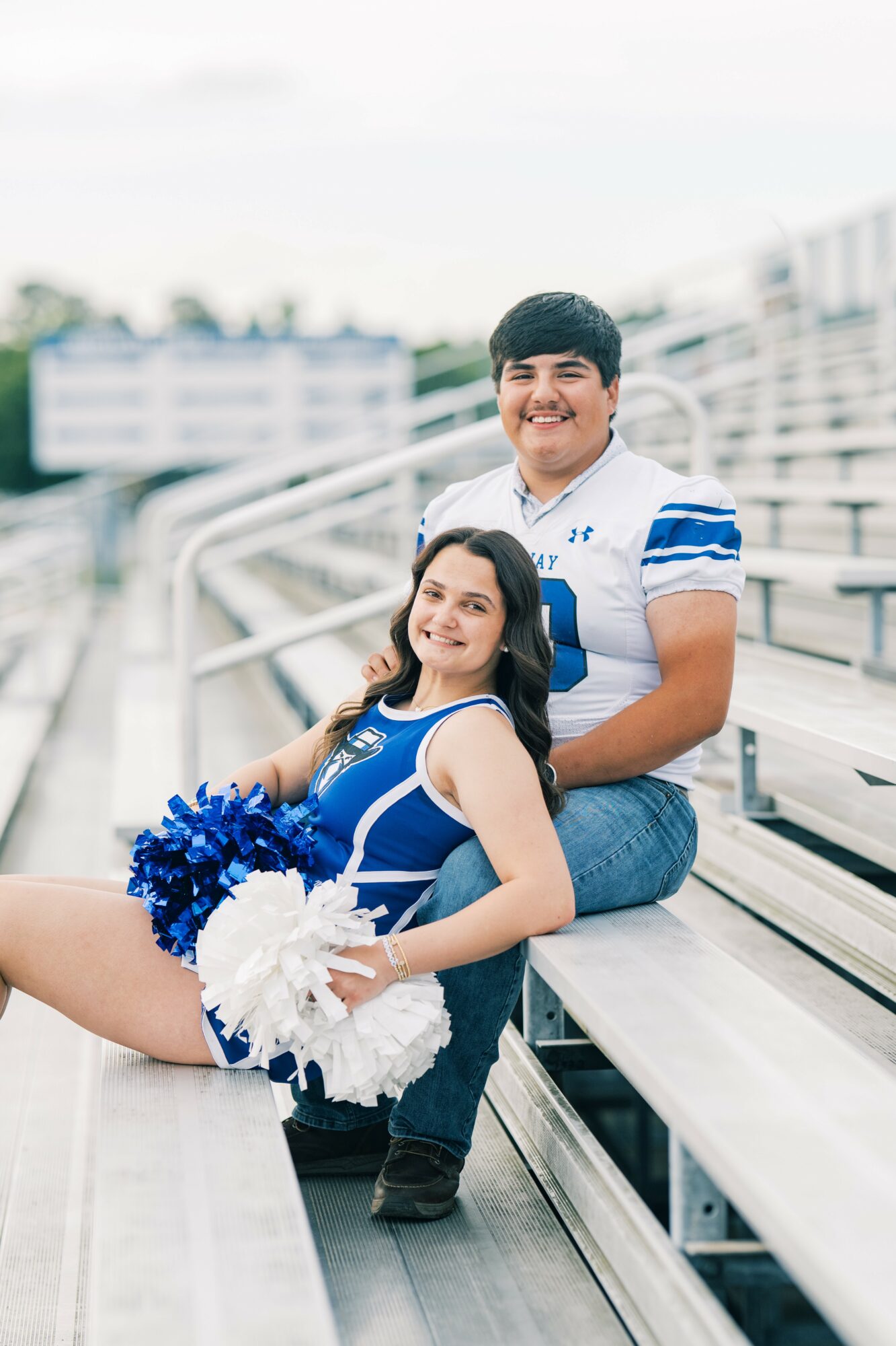 Two cheerleaders in blue and white uniforms sitting on stadium bleachers, smiling, with a blurred background of stadium and sky.