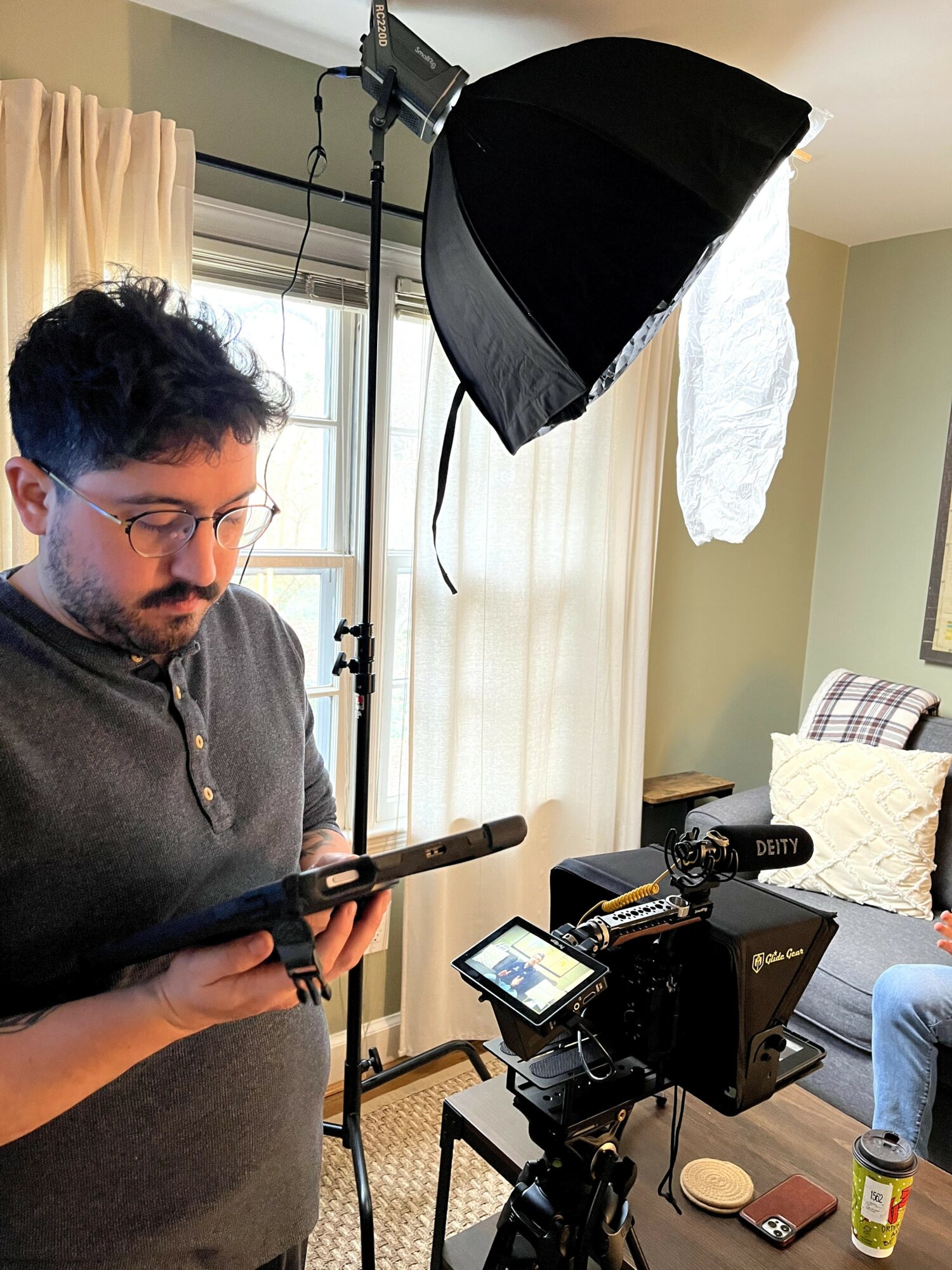 Man operating camera equipment in a room with a softbox light, window, and sofa, holding a remote control or monitor.