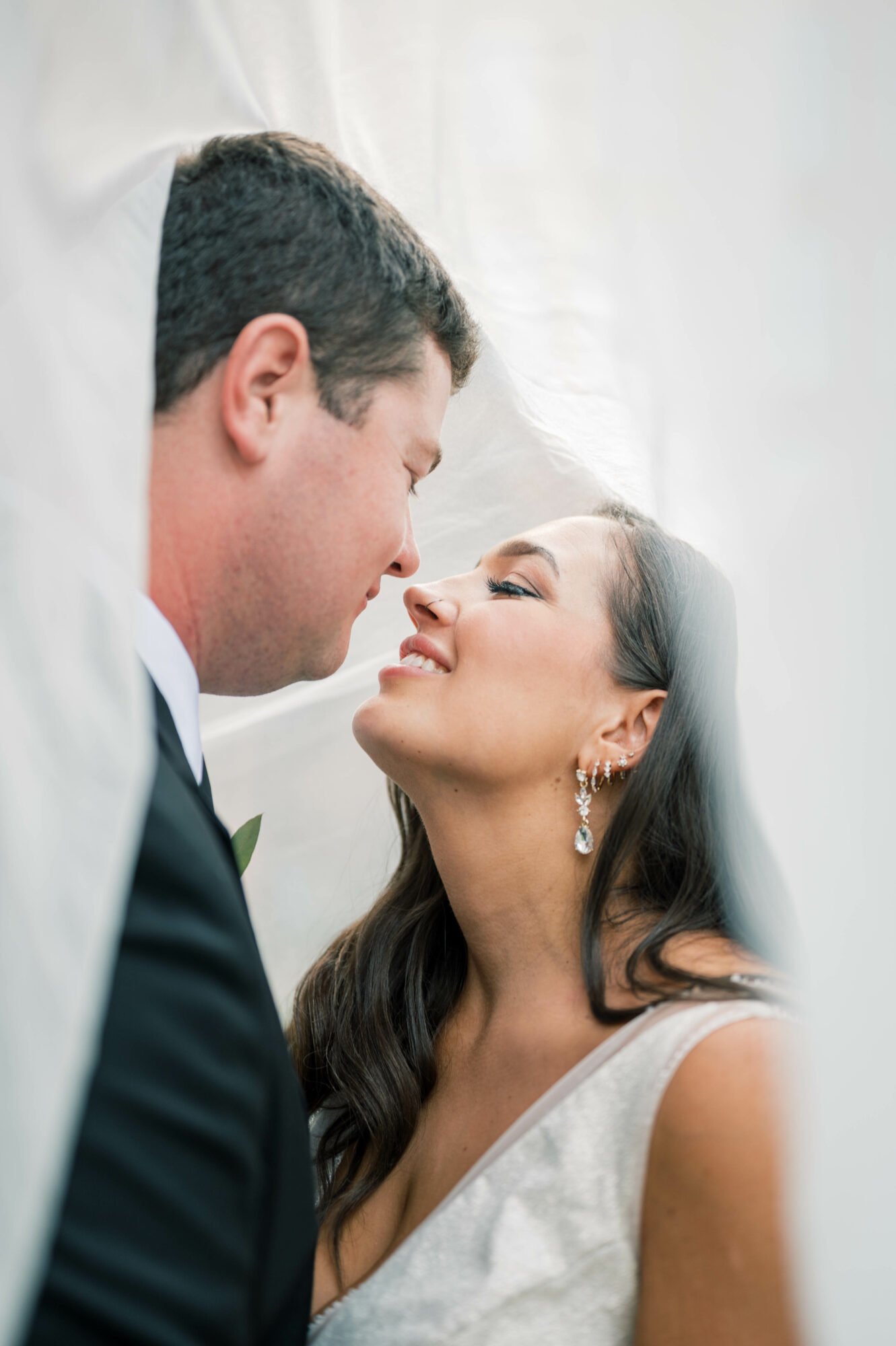 A bride and groom face each other closely, smiling with their eyes closed, under a white veil, in a wedding setting.