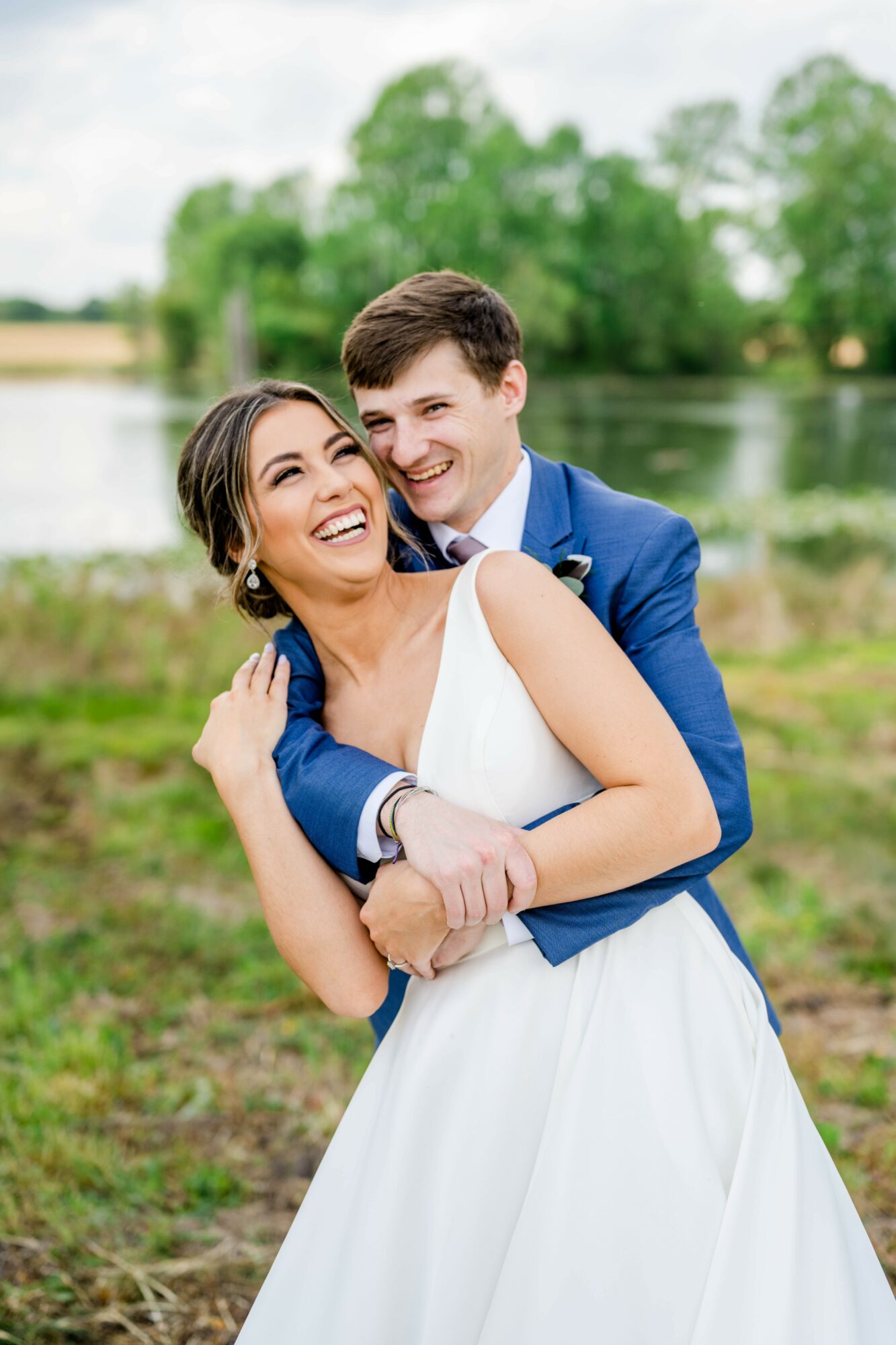 A smiling couple in wedding attire hugging outdoors near a lake with trees in the background.