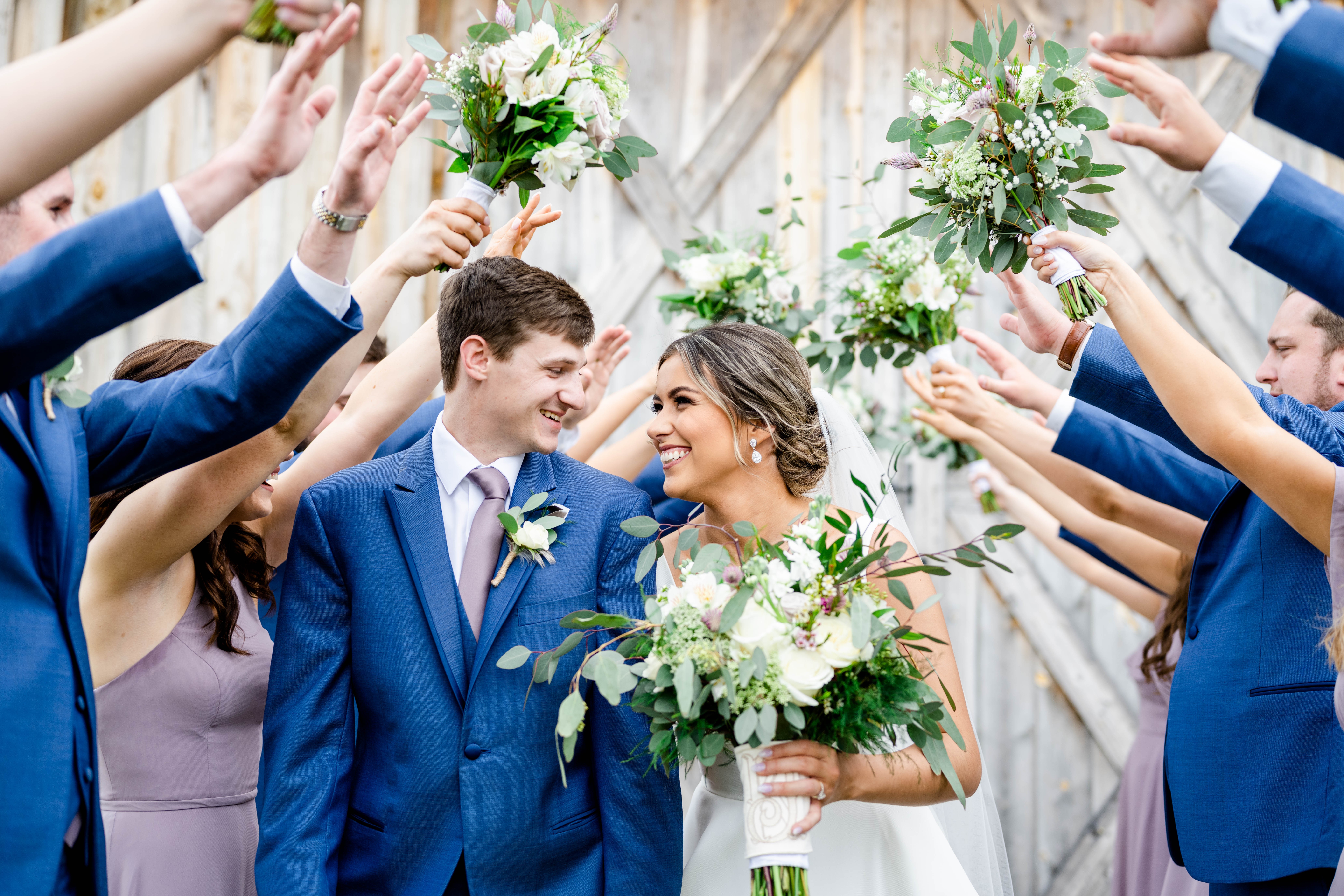 Bride and groom smiling, surrounded by friends raising bouquets in celebration, outdoors with wooden fence background.