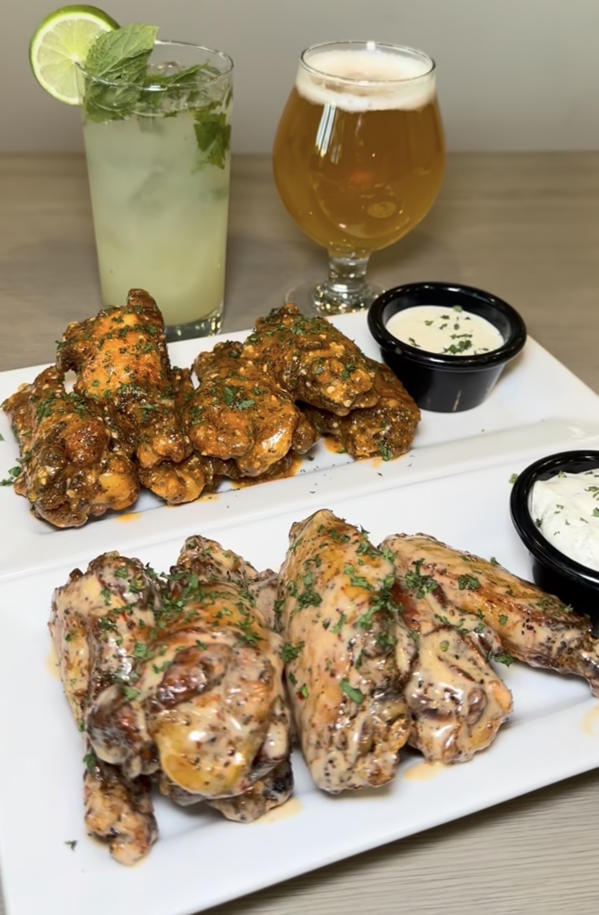 Two plates of chicken wings, a mojito, and a beer on a table with dipping sauces.